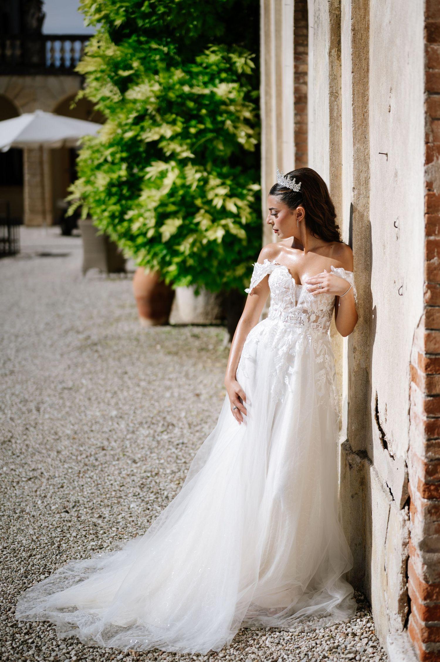 Bride in off-shoulder lace tulle wedding gown leans against rustic wall in elegant Italian villa courtyard.