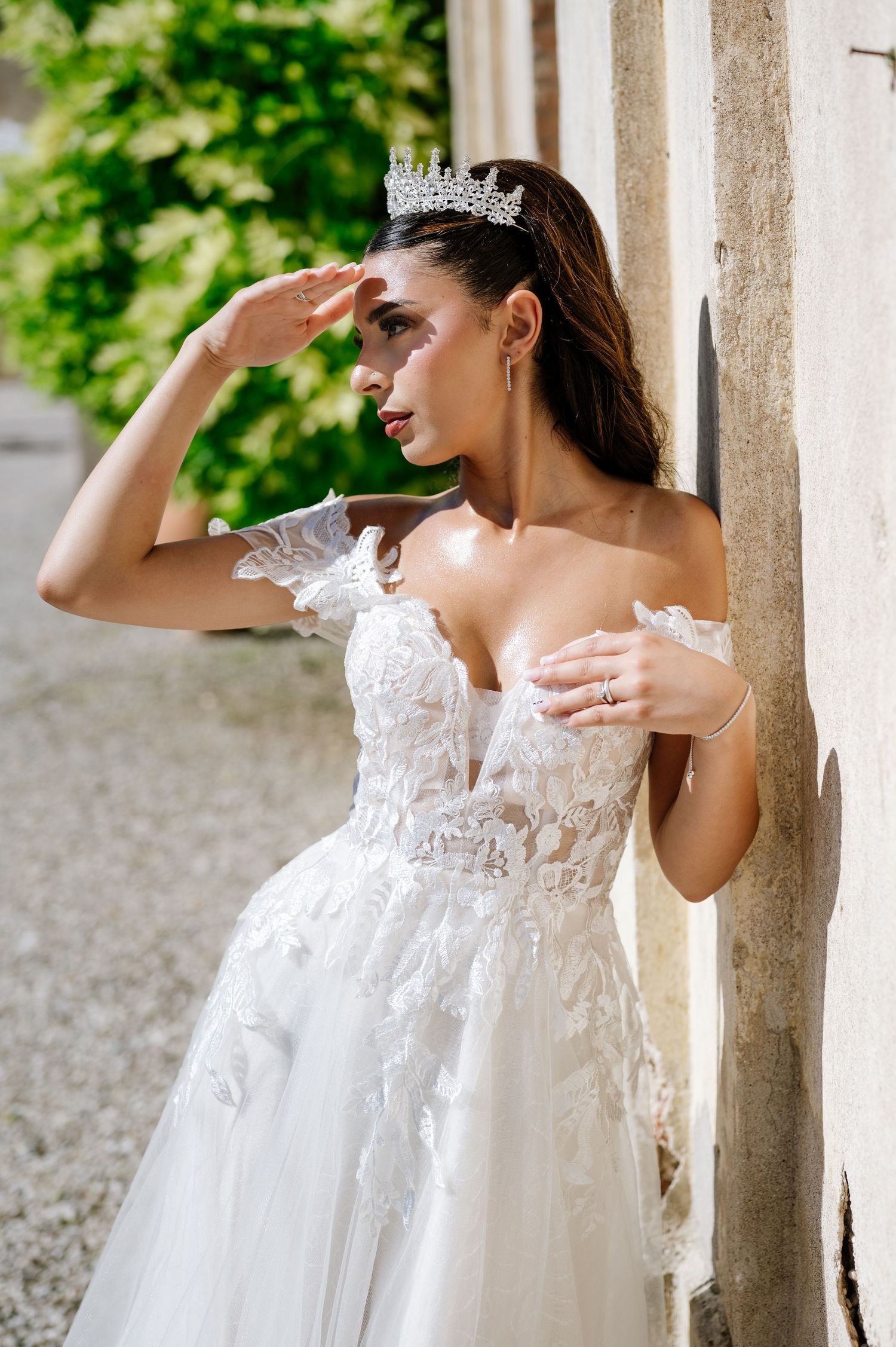 Bride in off-shoulder lace wedding gown and crystal tiara poses gracefully against a stone wall outdoors.