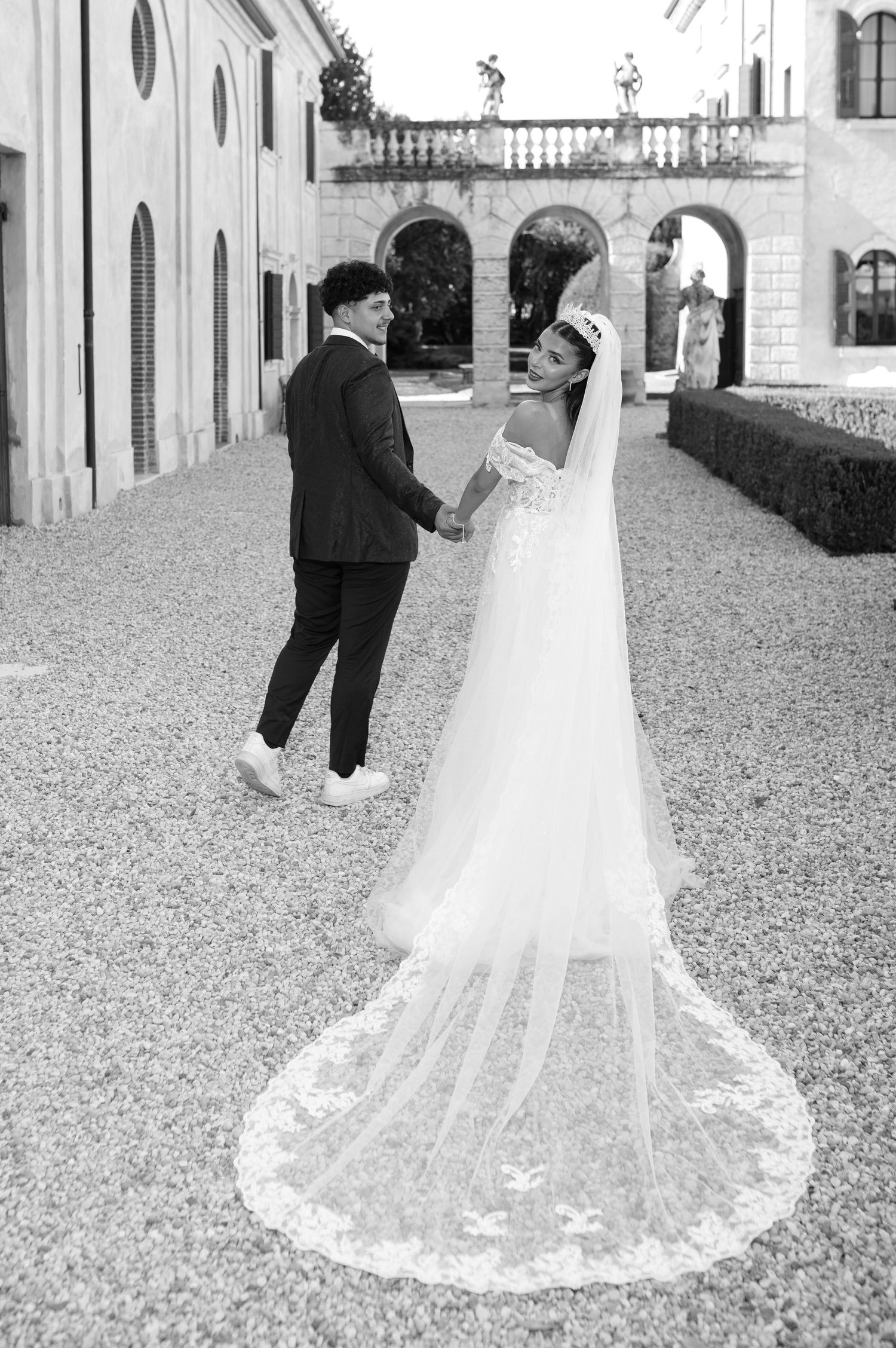 Black and white photo of a bride and groom holding hands at an elegant villa courtyard, her lace cathedral veil trailing gracefully.