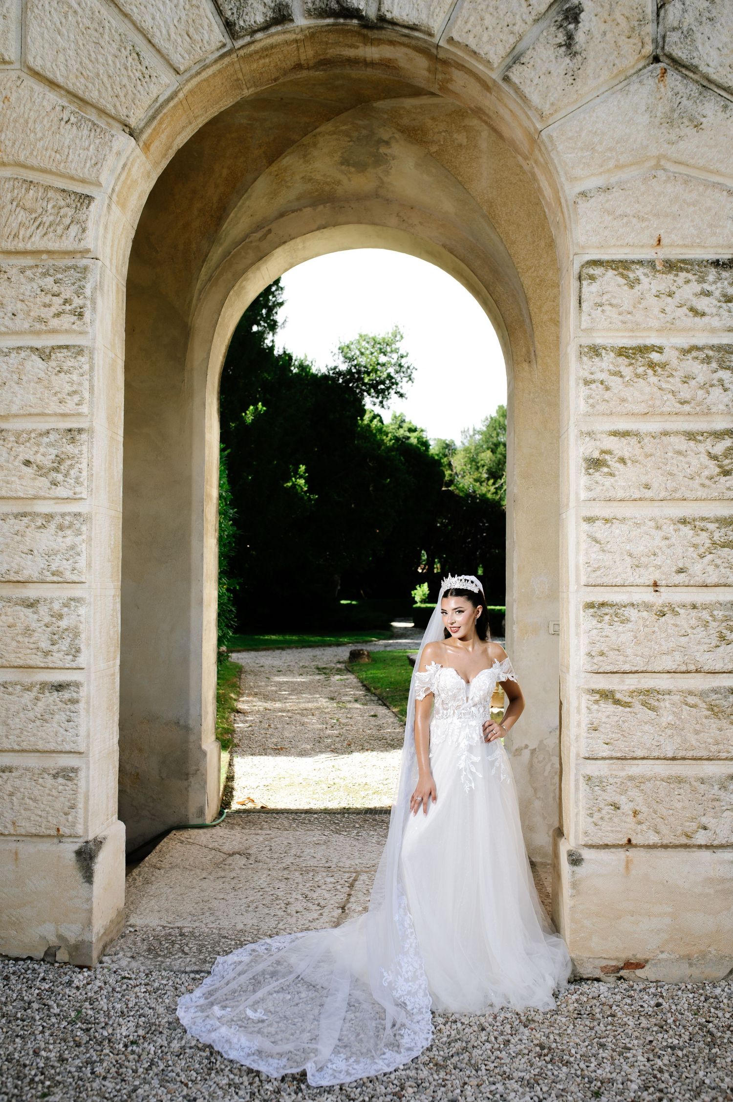 Bride in off-shoulder lace wedding gown with long train stands under stone arch in elegant garden setting.