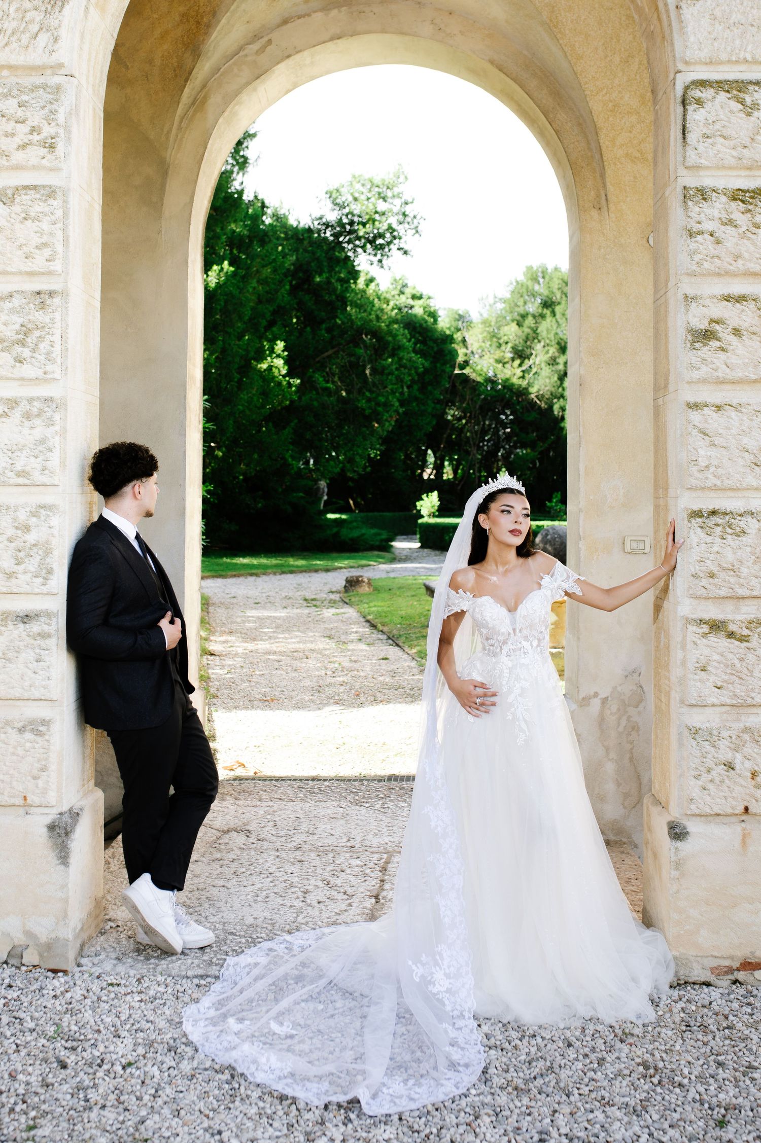 Bride in white lace gown and groom in black suit pose under stone arch in elegant outdoor wedding photo.