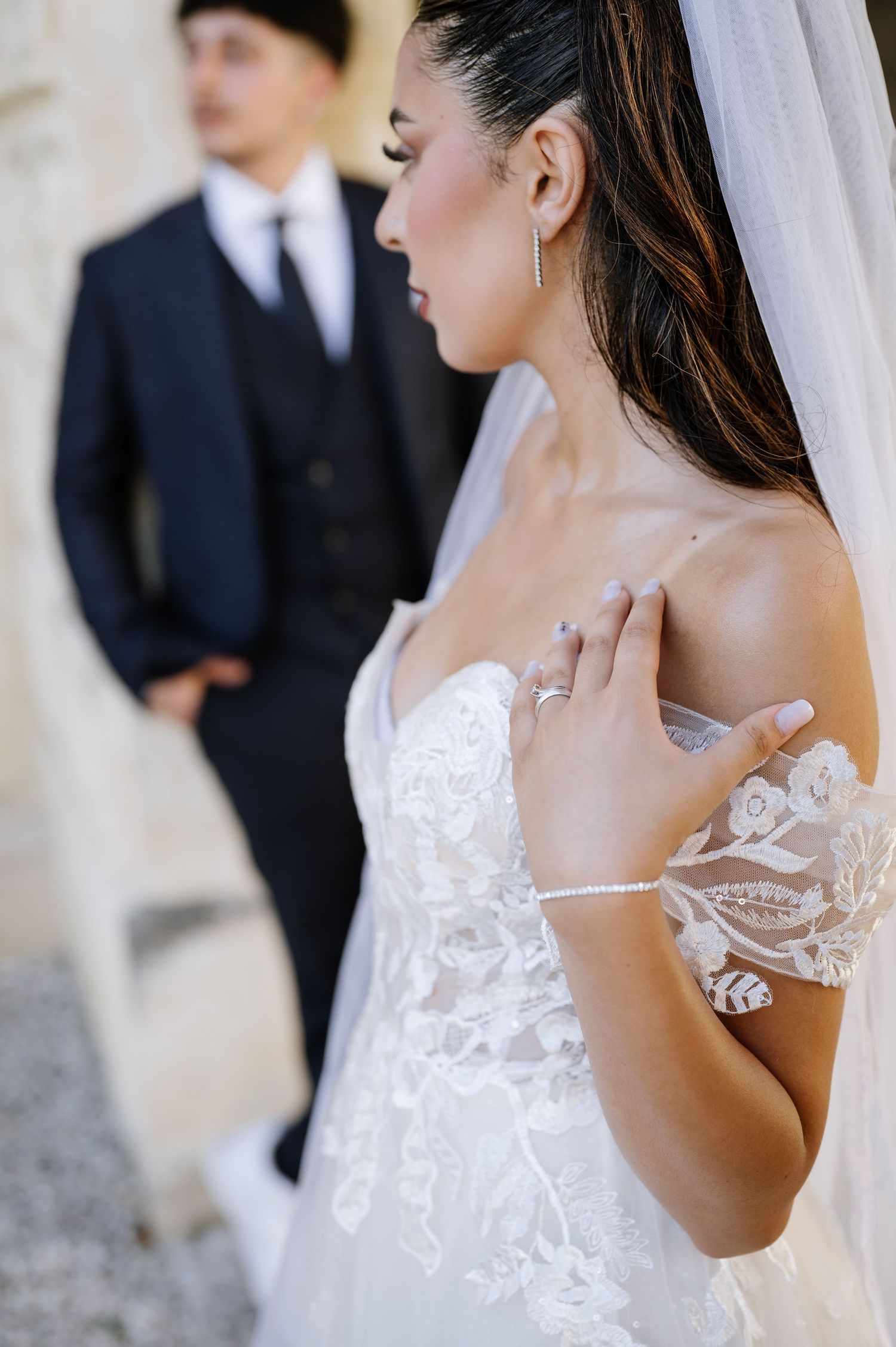 Close-up of bride in lace off-shoulder gown with veil and bracelet, groom in navy suit blurred behind.