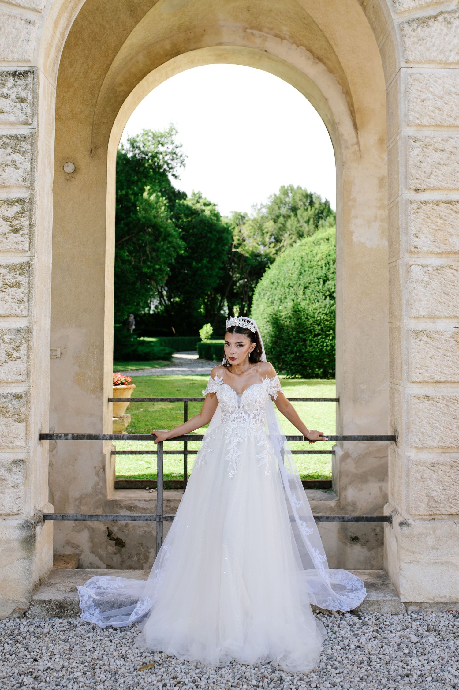 Bride in off-shoulder white lace ball gown and tiara stands gracefully under a stone arch in a garden.