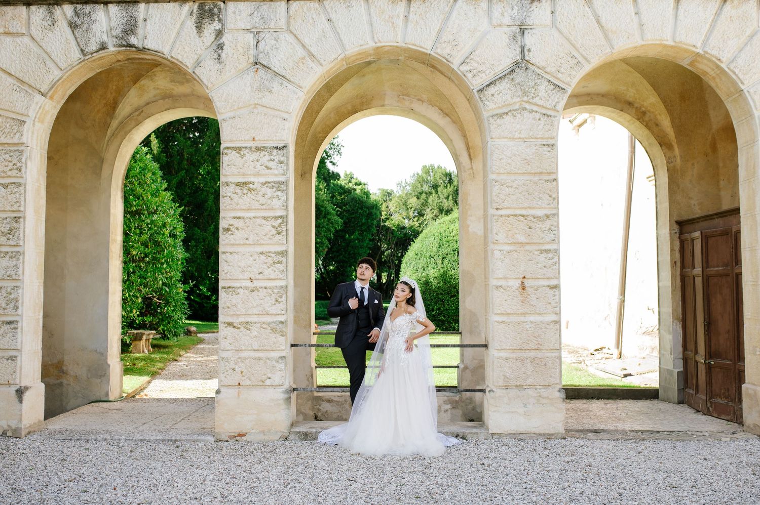 Bride and groom stand beneath stone arched colonnade of elegant historic venue on their wedding day.
