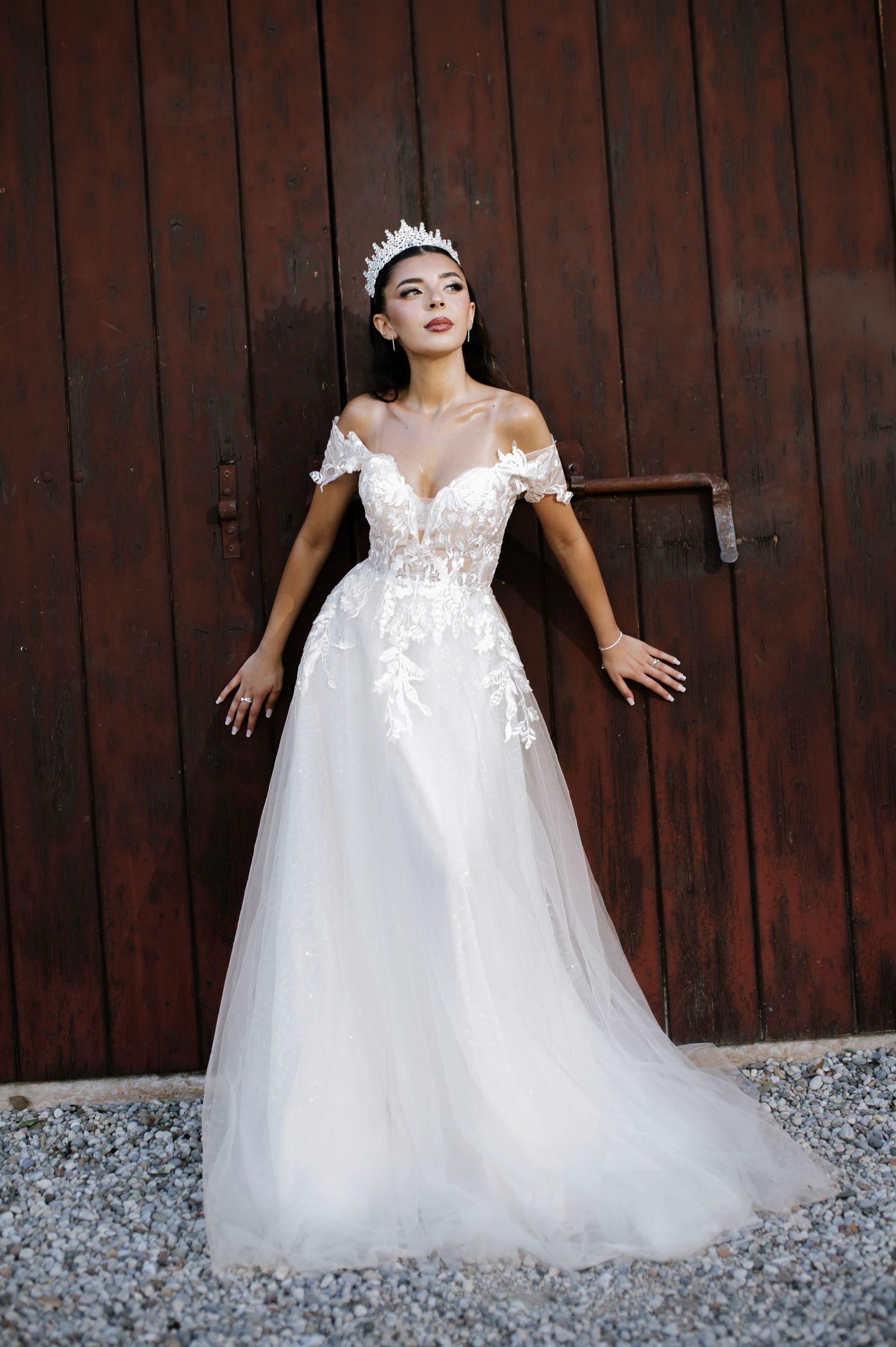 Bride in off-shoulder lace A-line wedding gown with floral appliques and crown poses by rustic wooden door.