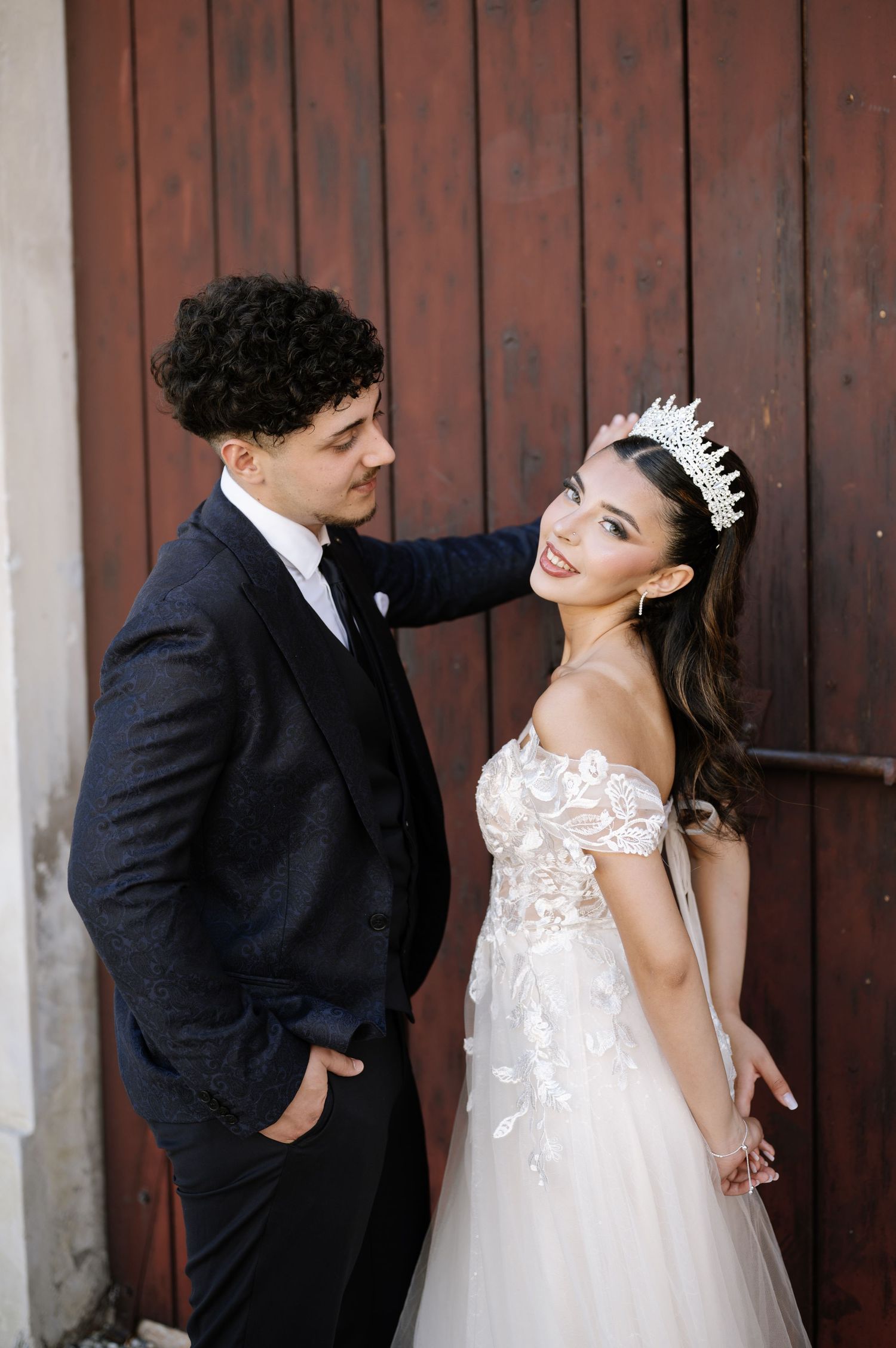 Bride in floral off-shoulder gown and tiara poses with groom in navy suit by rustic wooden door.