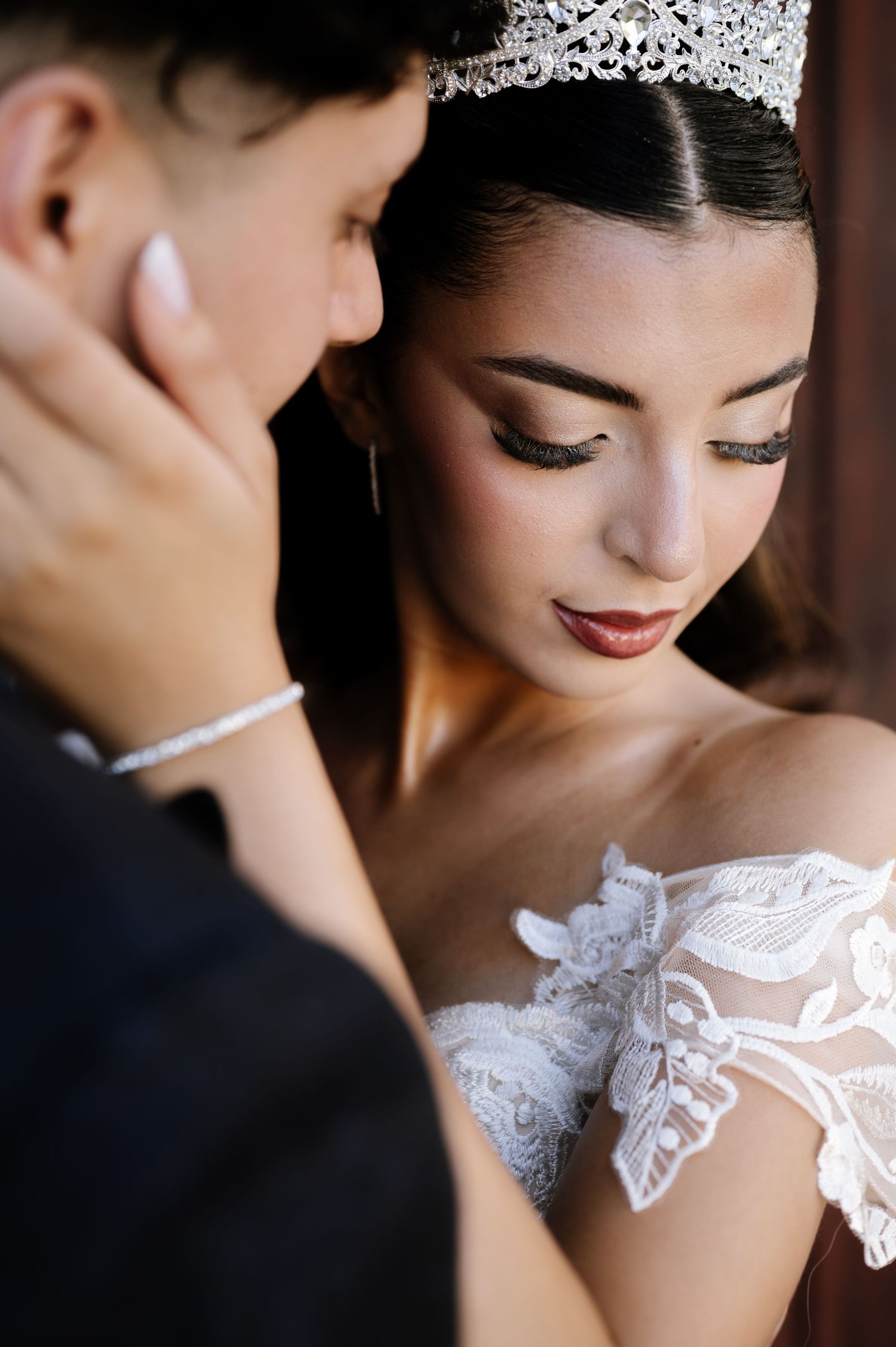 Bride in lace off-shoulder gown and crystal crown shares intimate moment with groom during wedding ceremony.