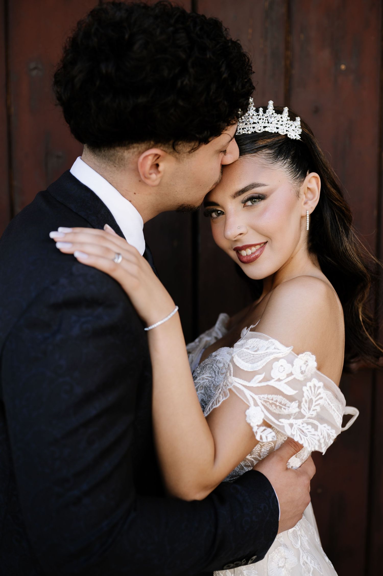Bride in lace off-shoulder gown and tiara embraces groom in black suit during romantic wedding portrait.