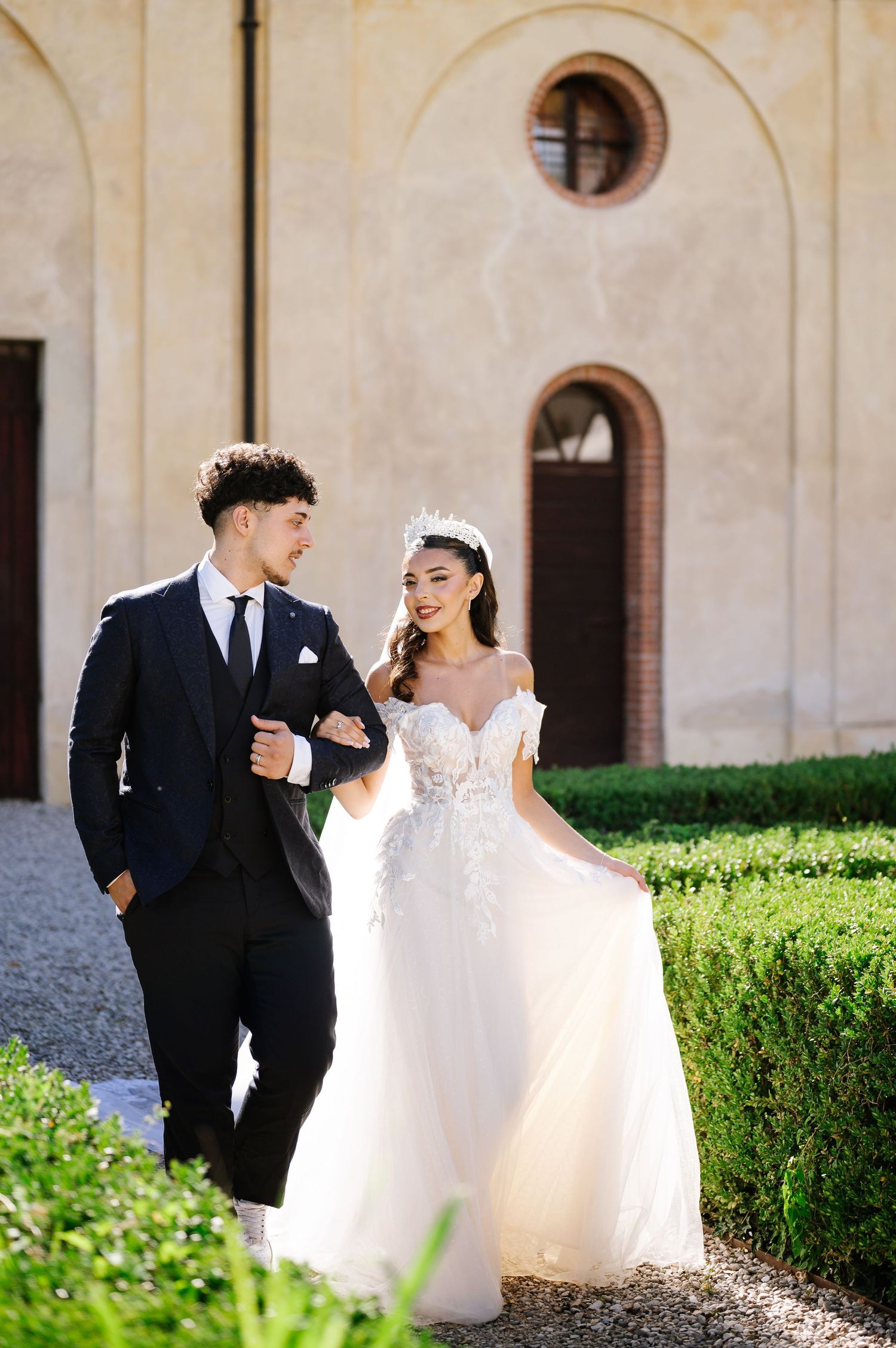 Elegant bride and groom walking outdoors at a sunlit Italian villa, wearing a white gown and navy suit.