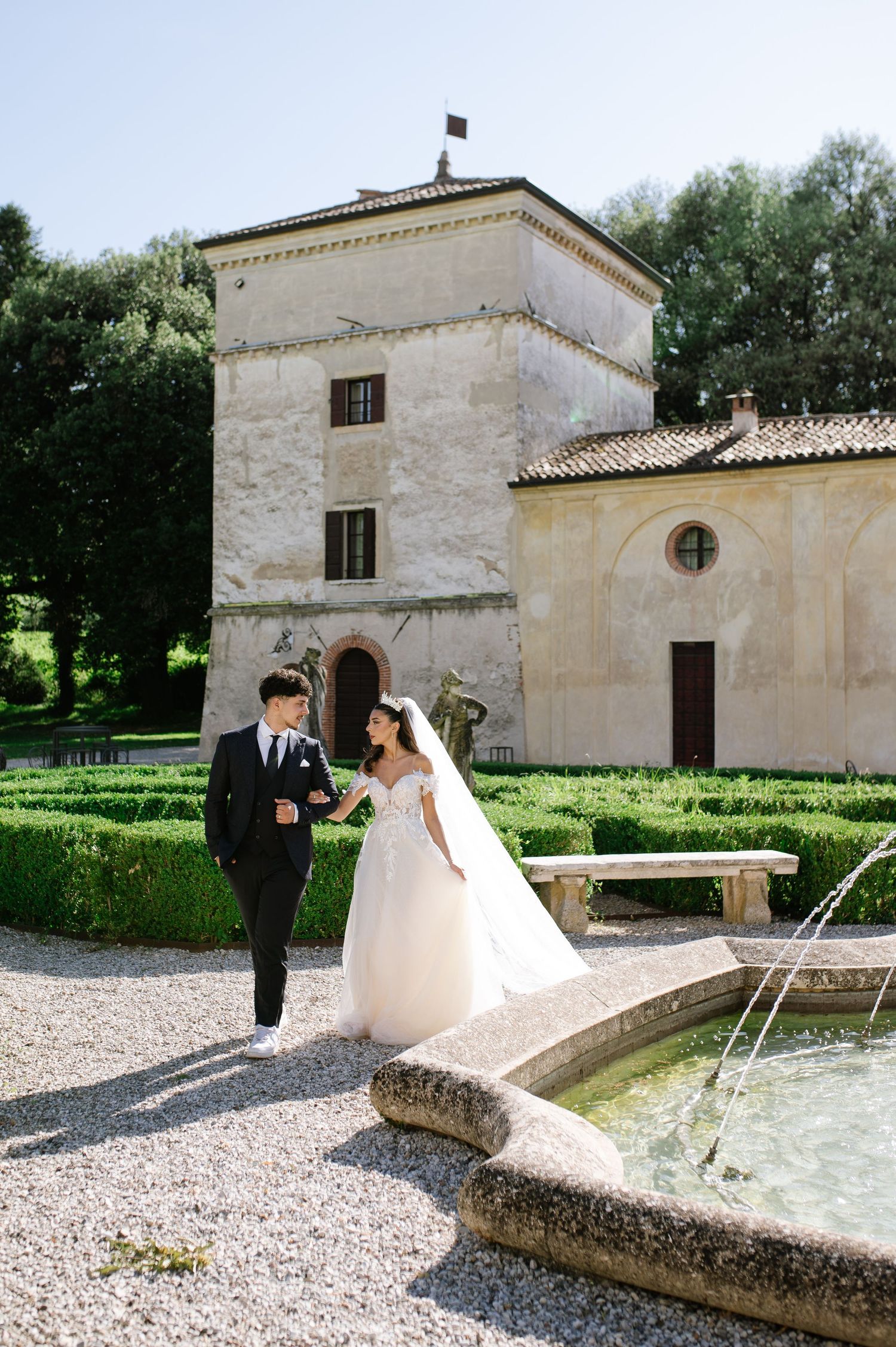 Wedding couple walks hand-in-hand by fountain at elegant Italian villa garden venue on sunny day.