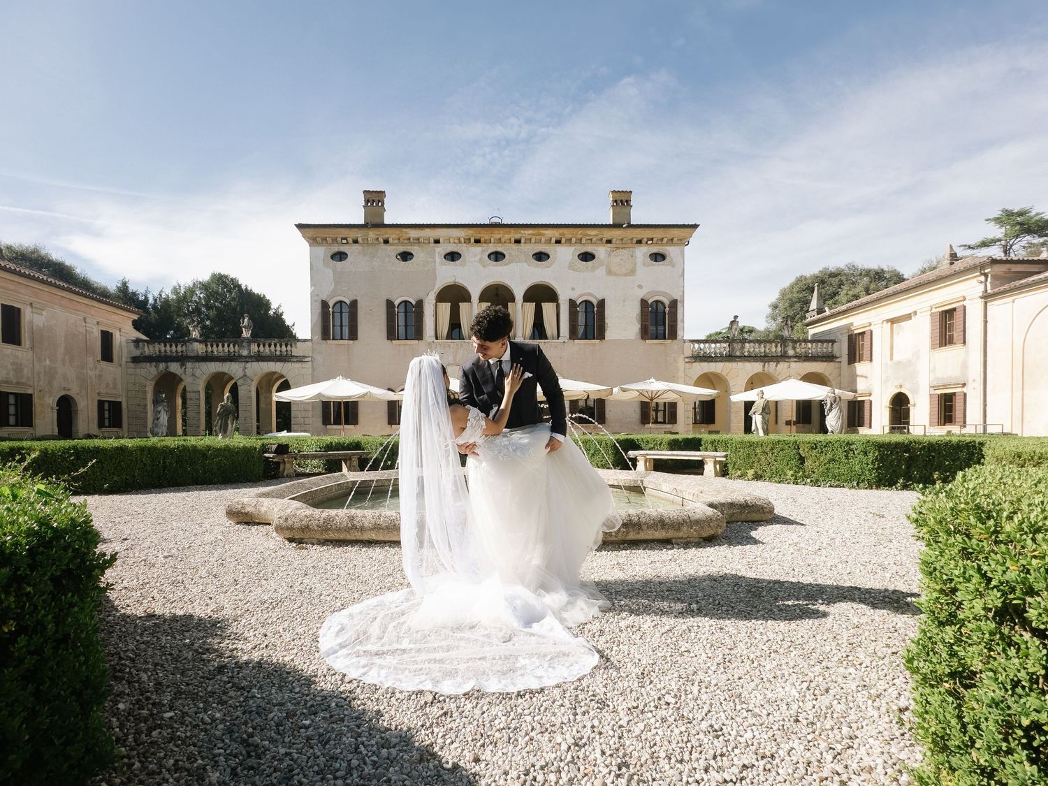 Bride and groom share a romantic kiss in front of an elegant Italian villa with manicured hedges and a fountain.