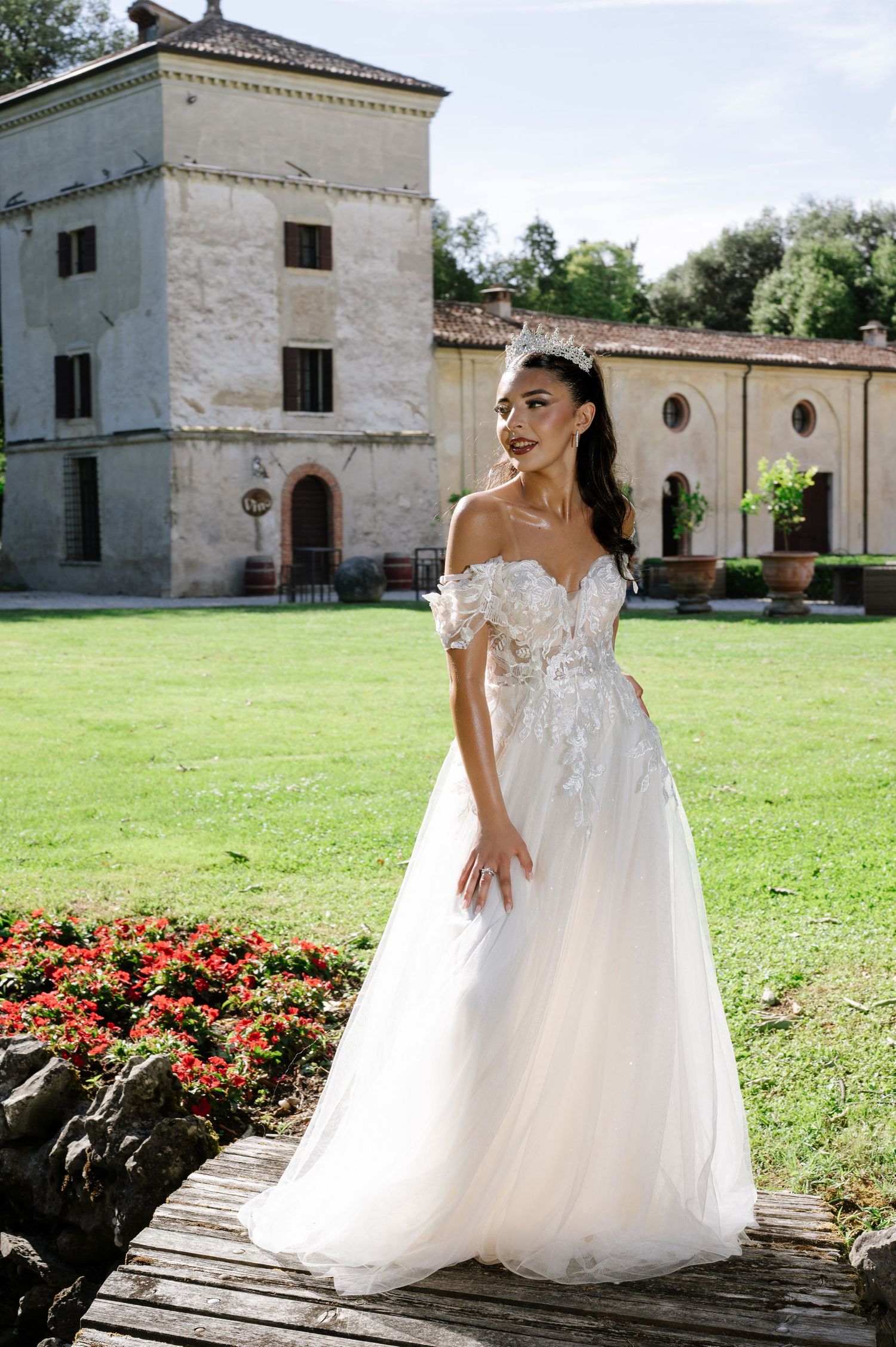 Bride in off-shoulder floral lace A-line wedding gown poses on wooden bridge before rustic Italian villa.