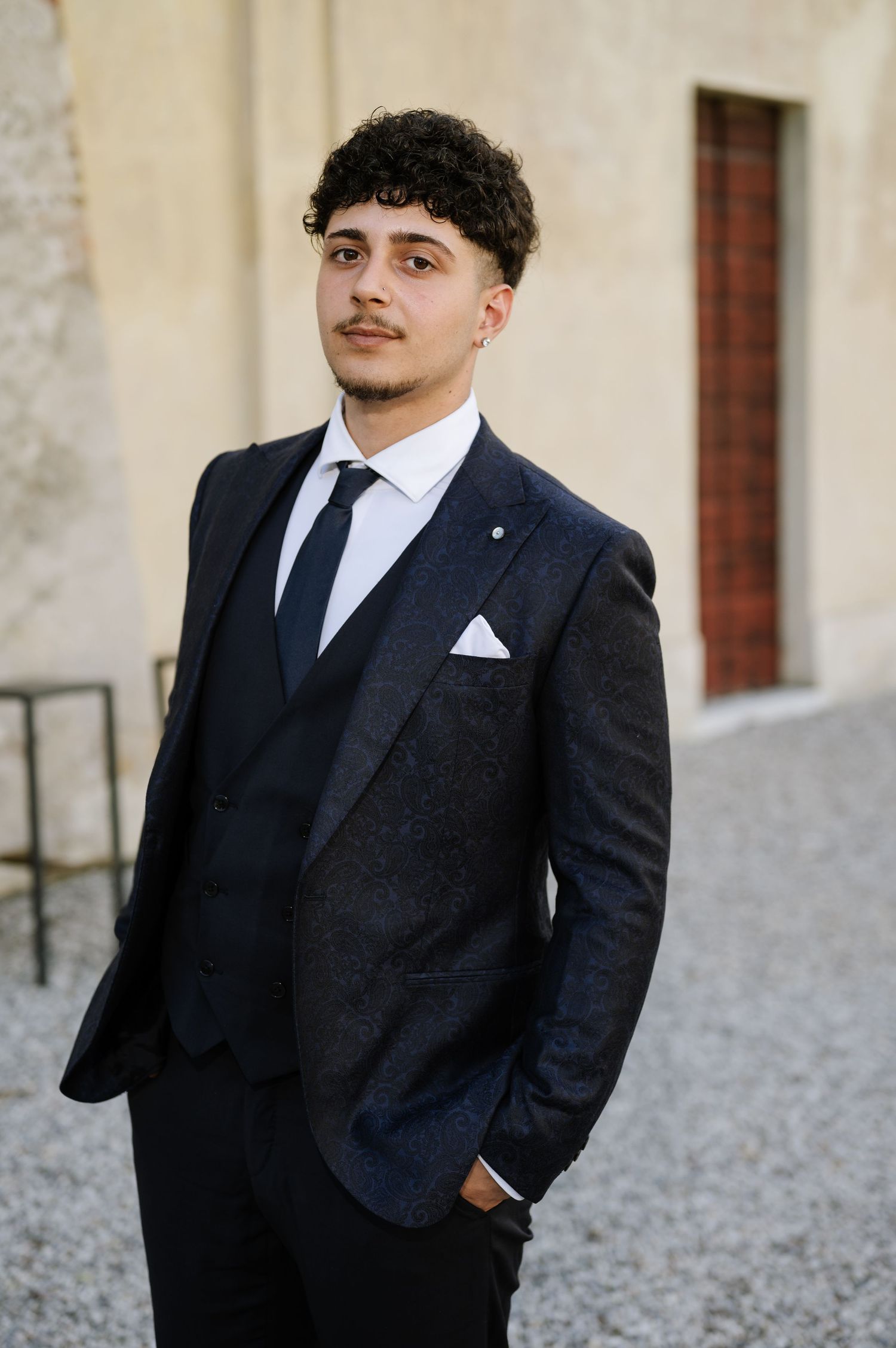 Young groom in tailored dark navy three-piece suit with white pocket square poses outdoors at wedding.