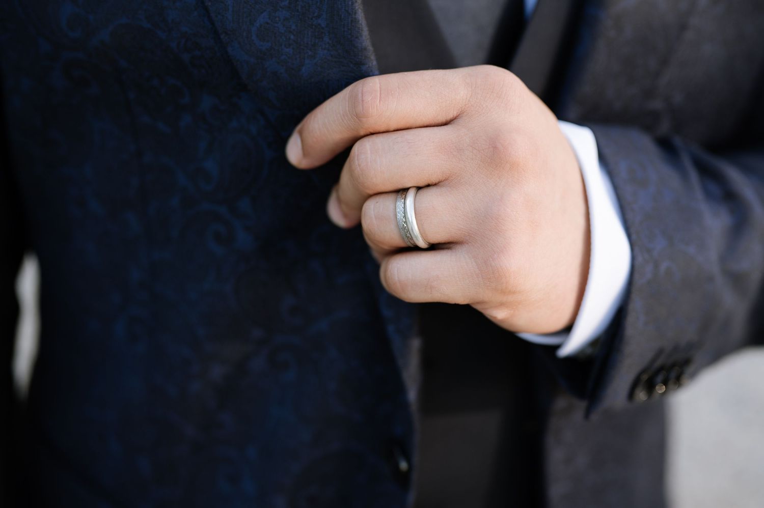 Close-up of groom's hand wearing a classic silver wedding band on ring finger in dark suit.