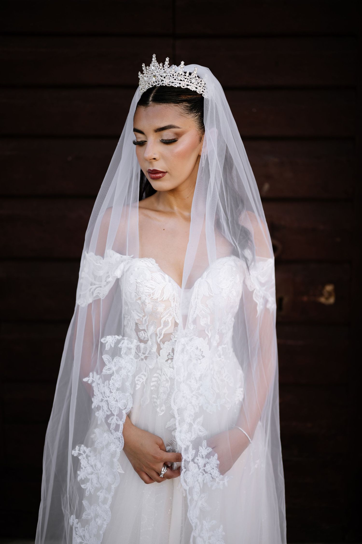 Bride wearing crystal tiara and lace veil in off-shoulder white wedding dress poses against dark wooden backdrop.