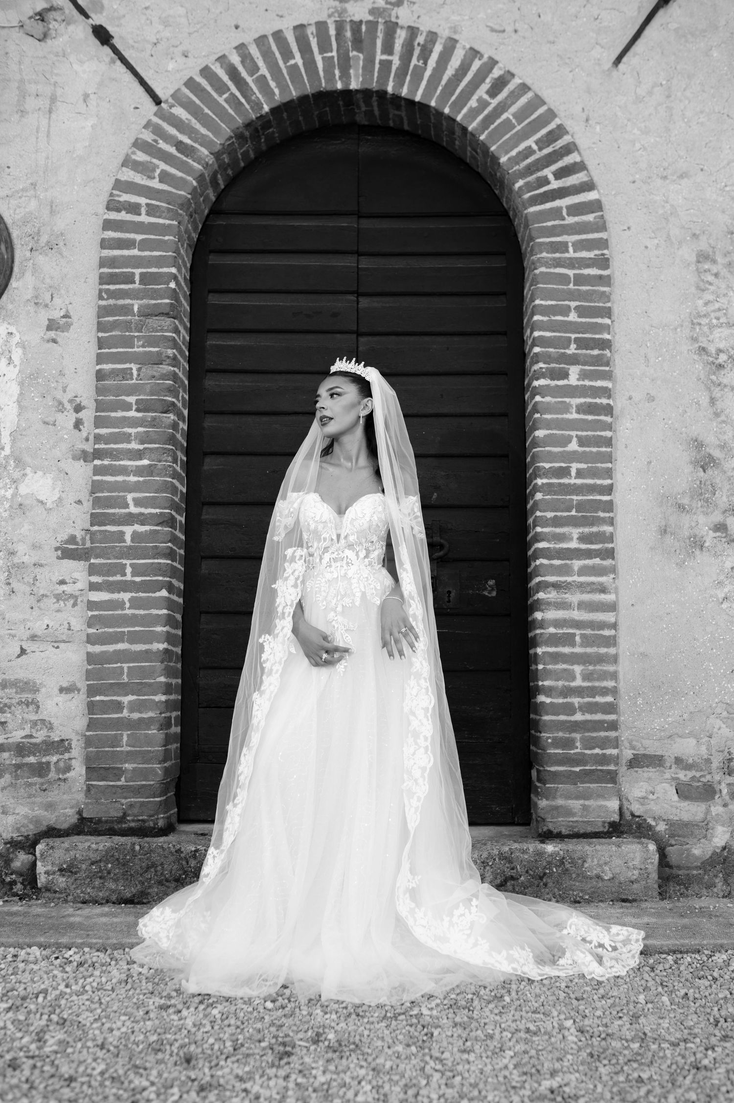 Elegant bride in lace ball gown with cathedral veil and crown poses before rustic arched doorway.