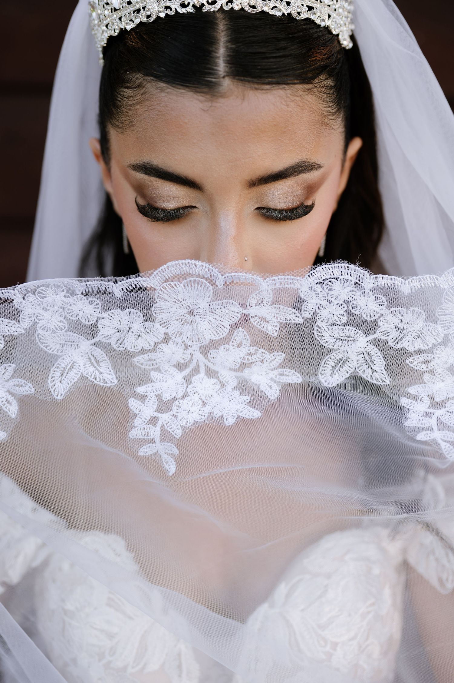 Close-up of bride holding embroidered lace veil over face, wearing tiara and dramatic eye makeup.