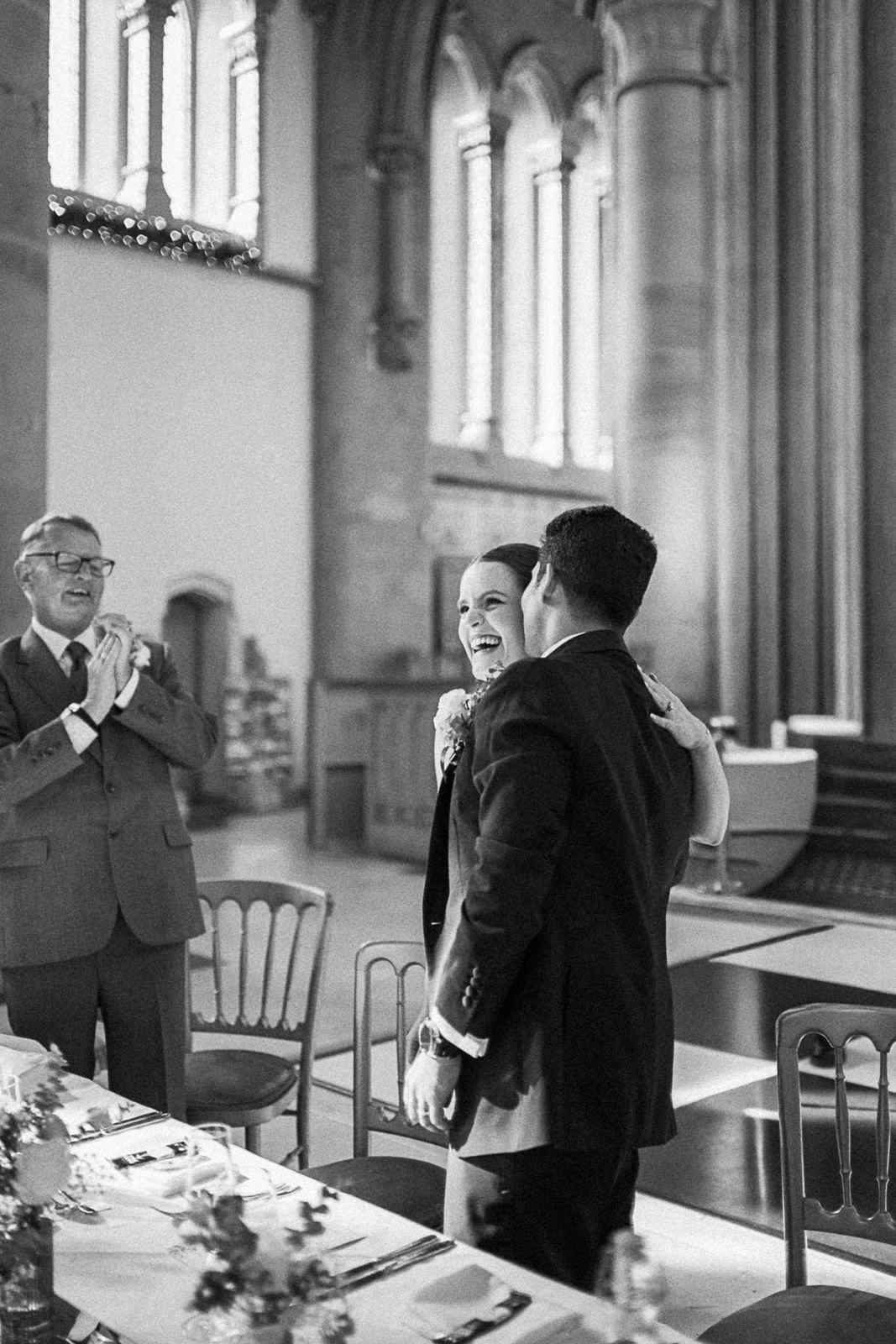 Black and white wedding photo of a joyful couple embracing in a grand church hall, with a guest applauding nearby.