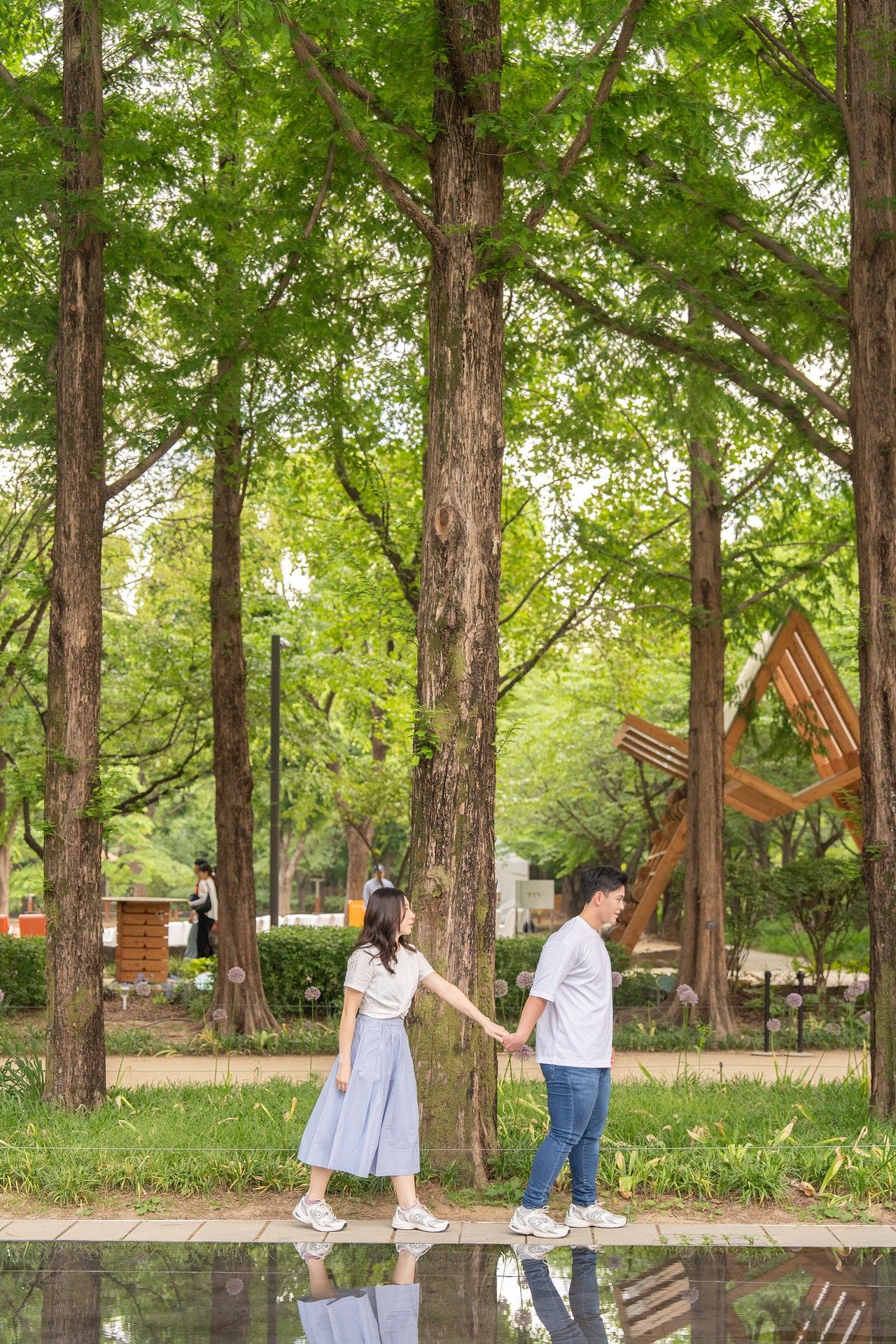 A couple holds hands while walking through a lush green park with tall trees and a reflective pool.