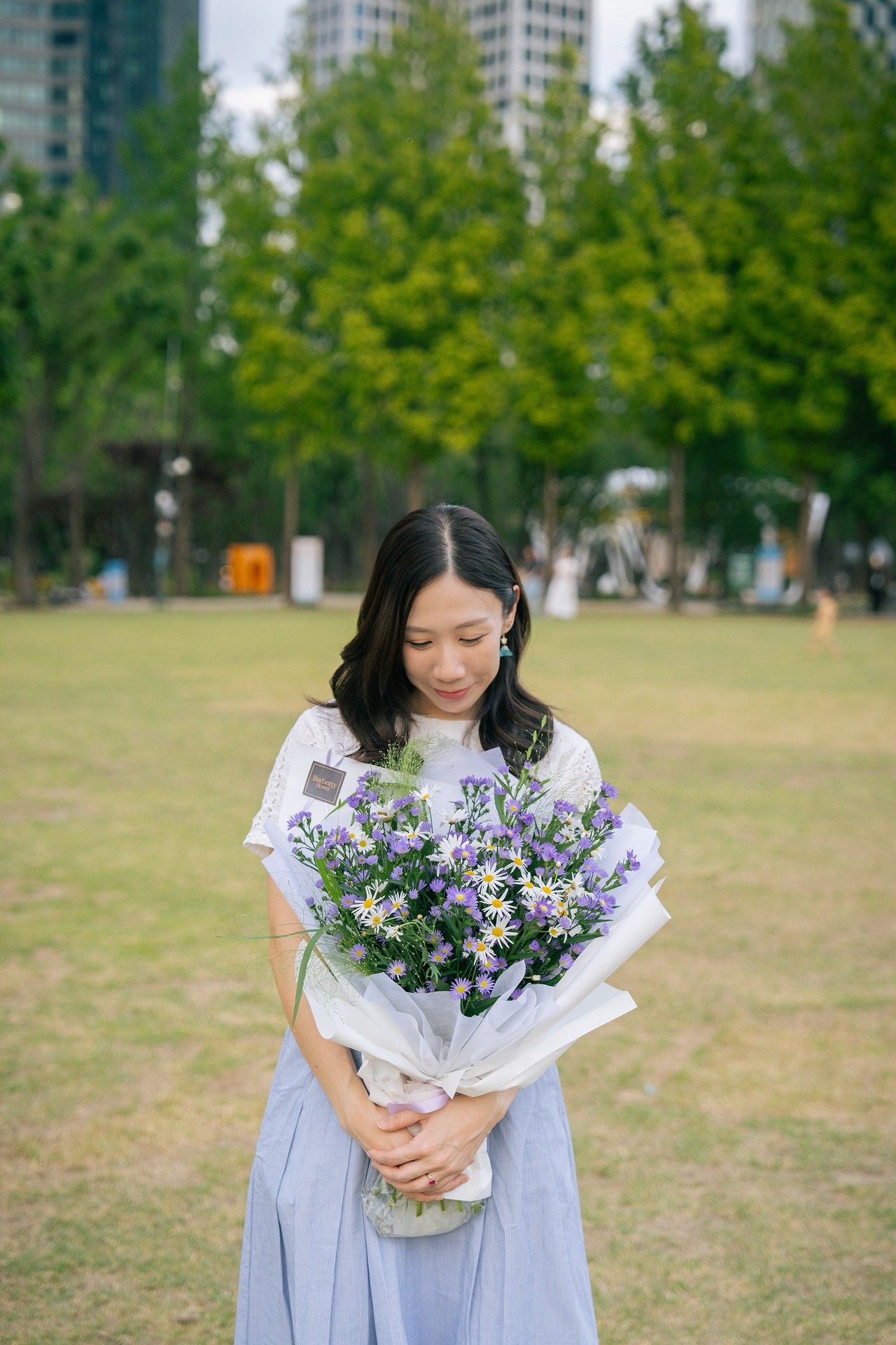 Young Asian woman in white top and blue skirt holding purple and white flower bouquet in a green park.