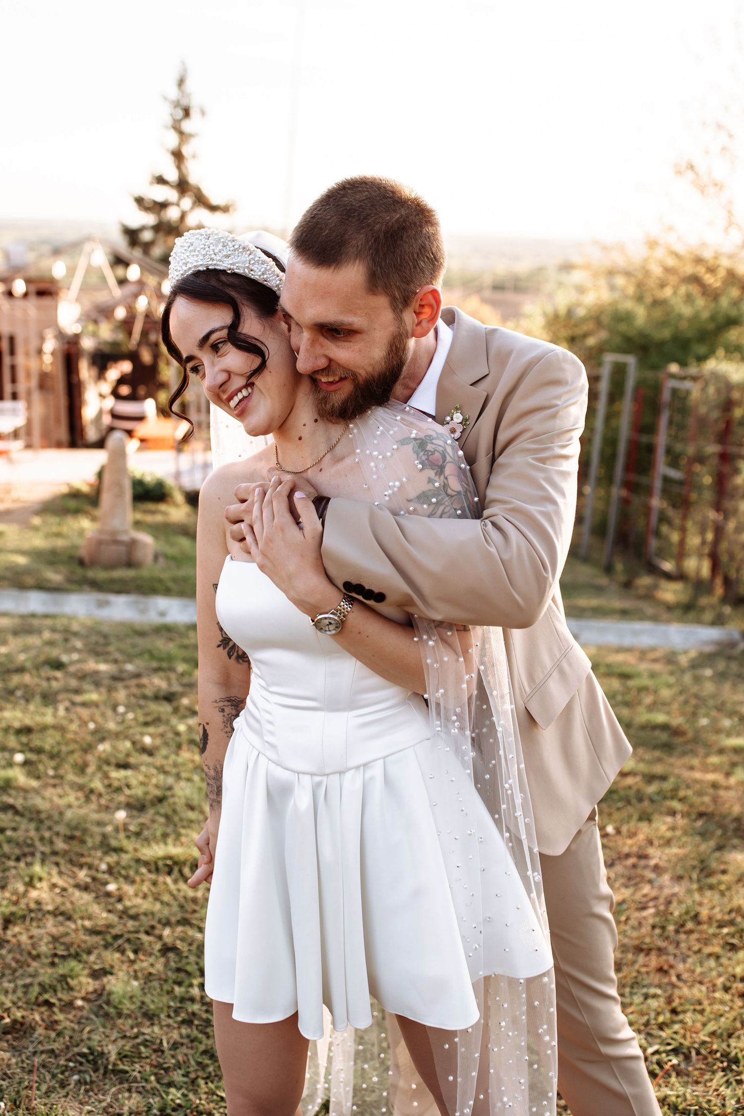 Esküvői kreatív fotózás. Happy bride in white mini dress and groom in beige suit embracing outdoors during golden hour wedding celebration.