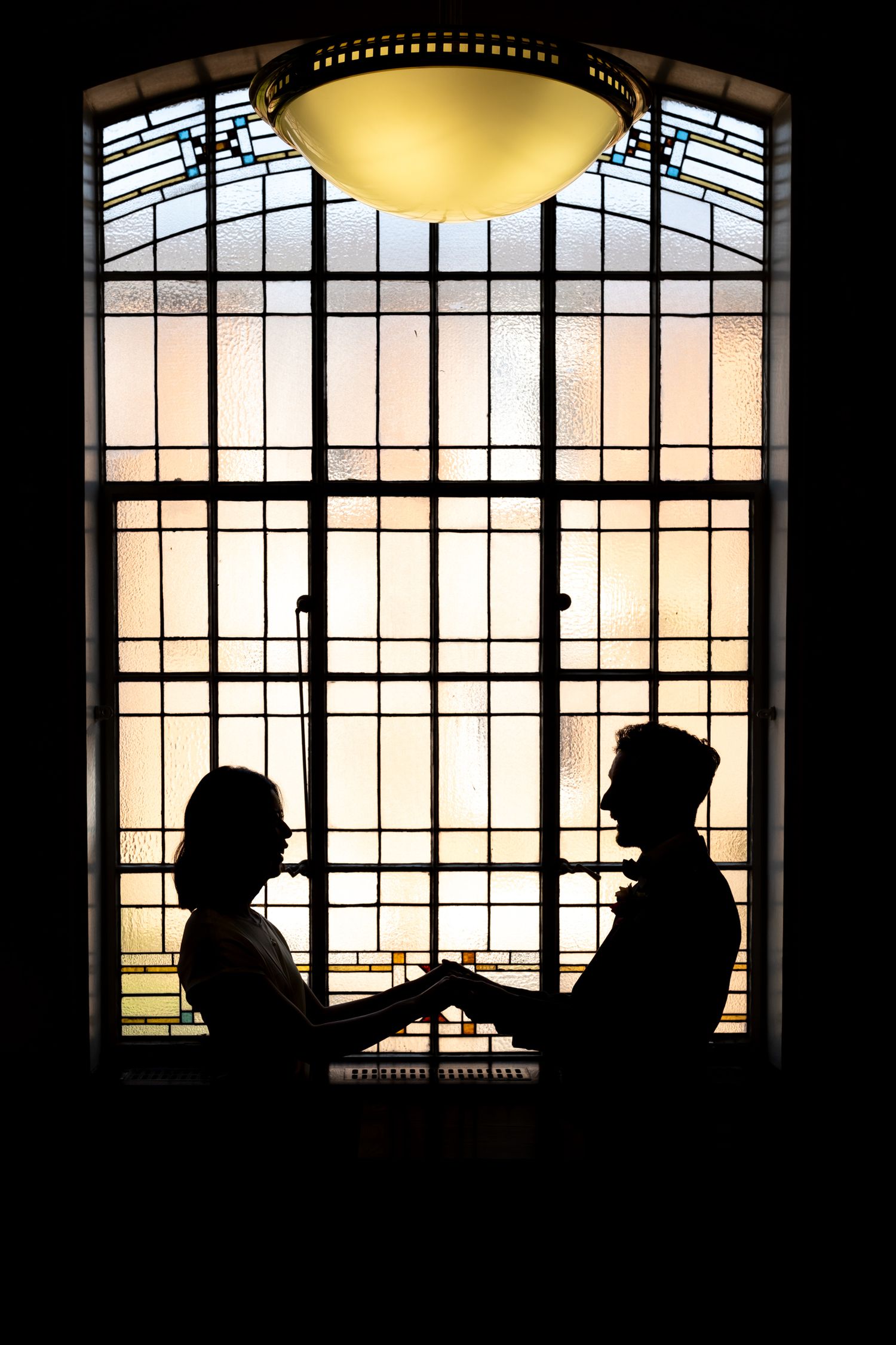 Romantic silhouette of couple holding hands in front of ornate stained glass window with warm backlight.