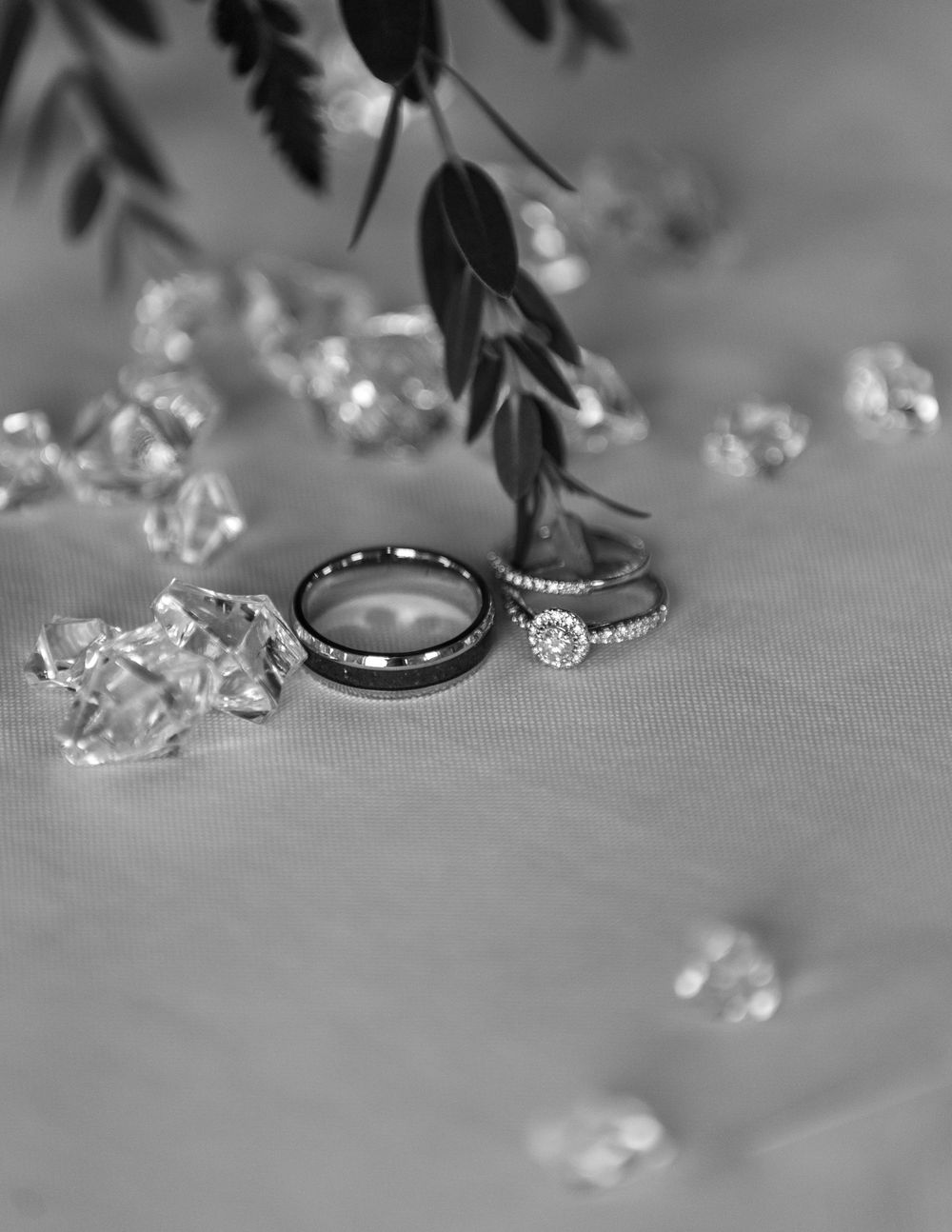 Black and white photo of wedding rings with diamond accents surrounded by crystal gems and greenery on soft fabric.