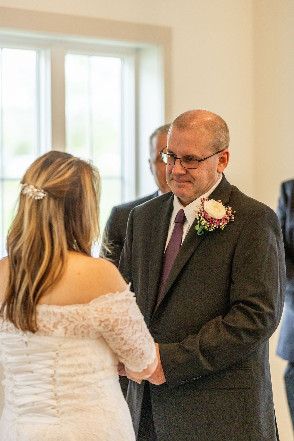 Father of the bride smiling emotionally while holding hands with bride in white lace dress during wedding ceremony.