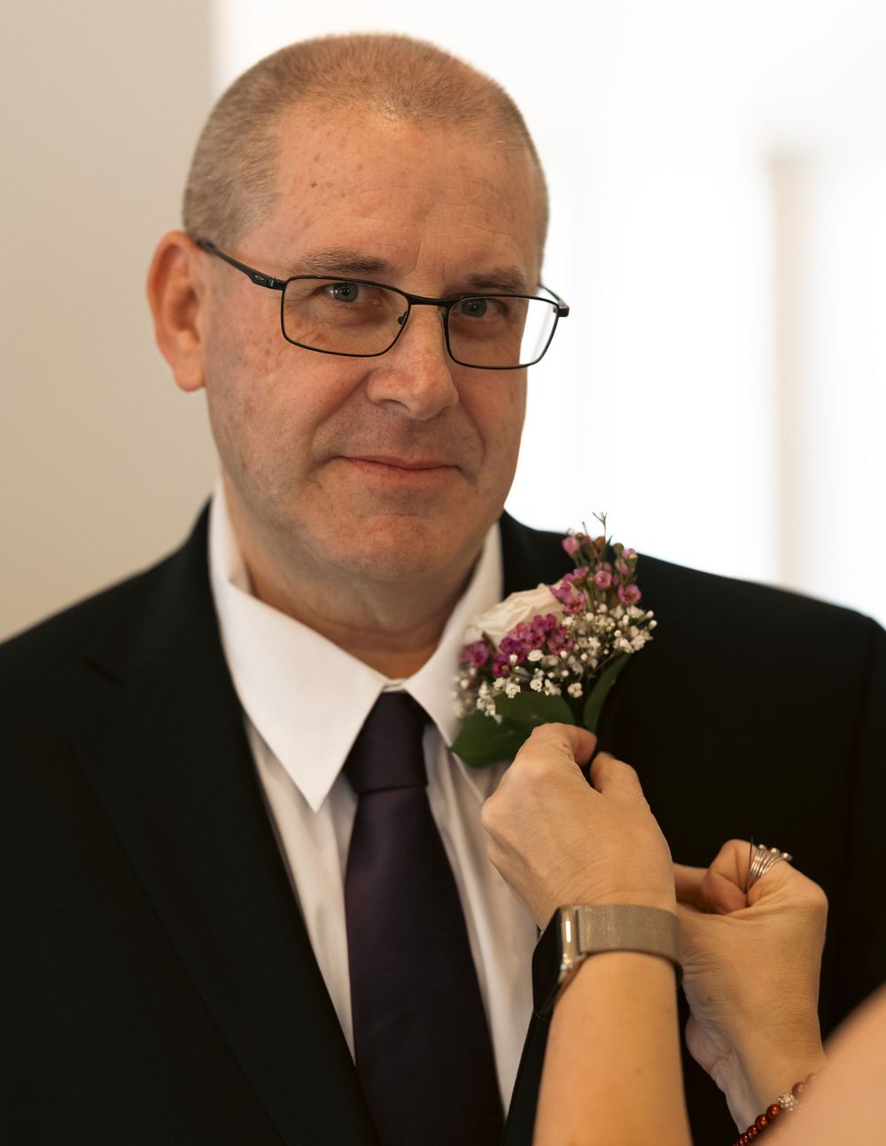 Man in black suit with purple tie and glasses having a floral boutonniere pinned to his lapel at a wedding.