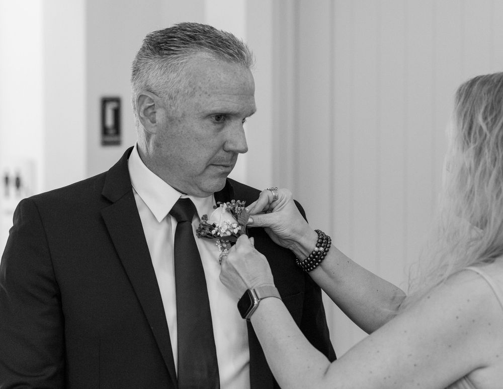Black and white photo of a man in a suit having a boutonniere pinned to his lapel at a wedding.