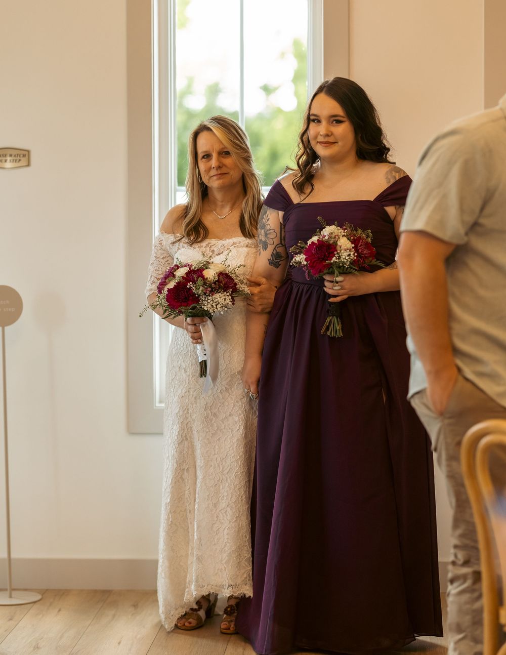 Bride in white lace dress and bridesmaid in purple gown holding red floral bouquets at wedding ceremony.
