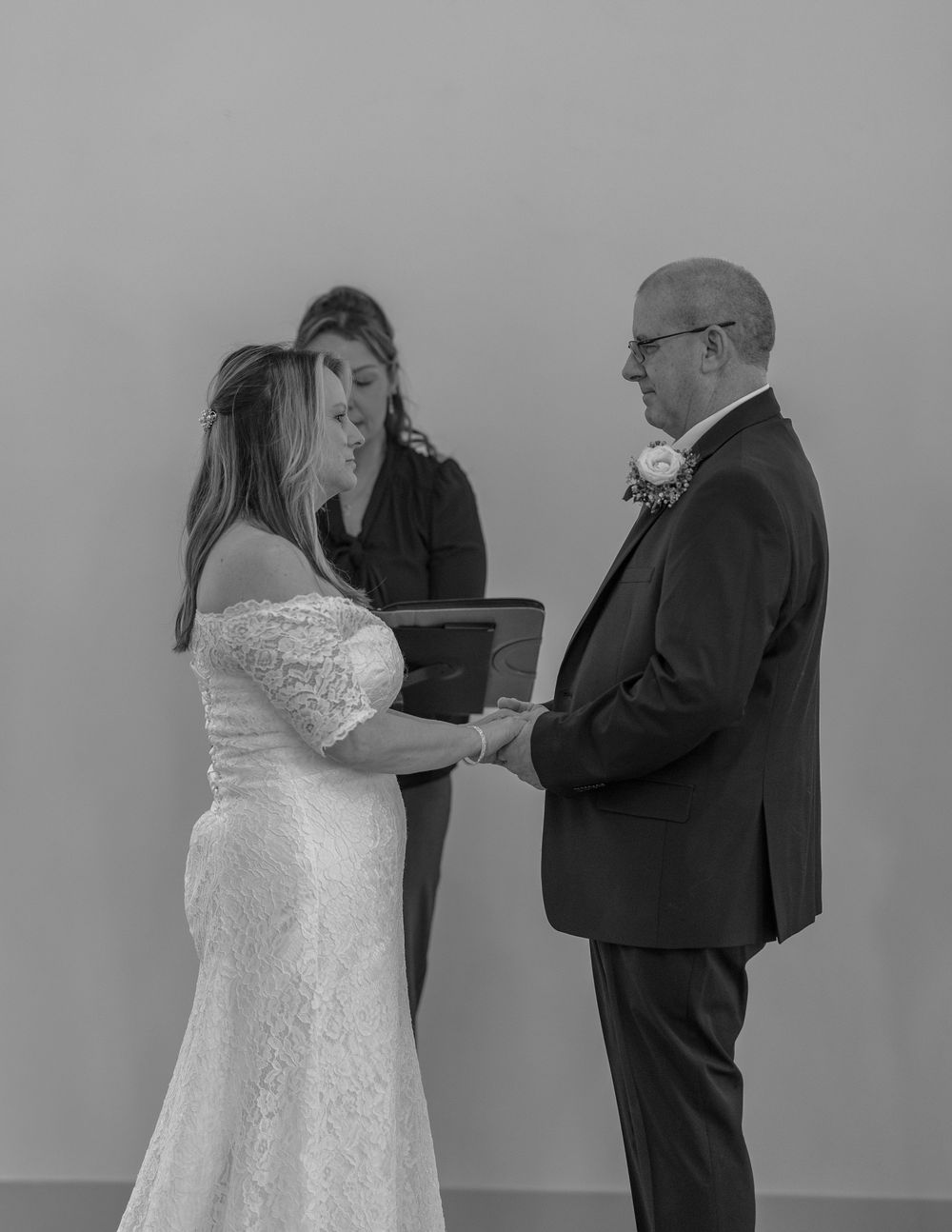 Black and white wedding ceremony photo of bride and groom exchanging vows with officiant holding book.