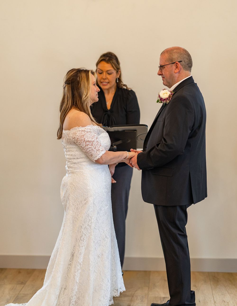 Couple exchanging wedding vows with smiling officiant holding book during intimate indoor ceremony.