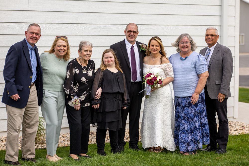 Group of eight people posing together outdoors at a wedding, with bride in white dress holding bouquet.