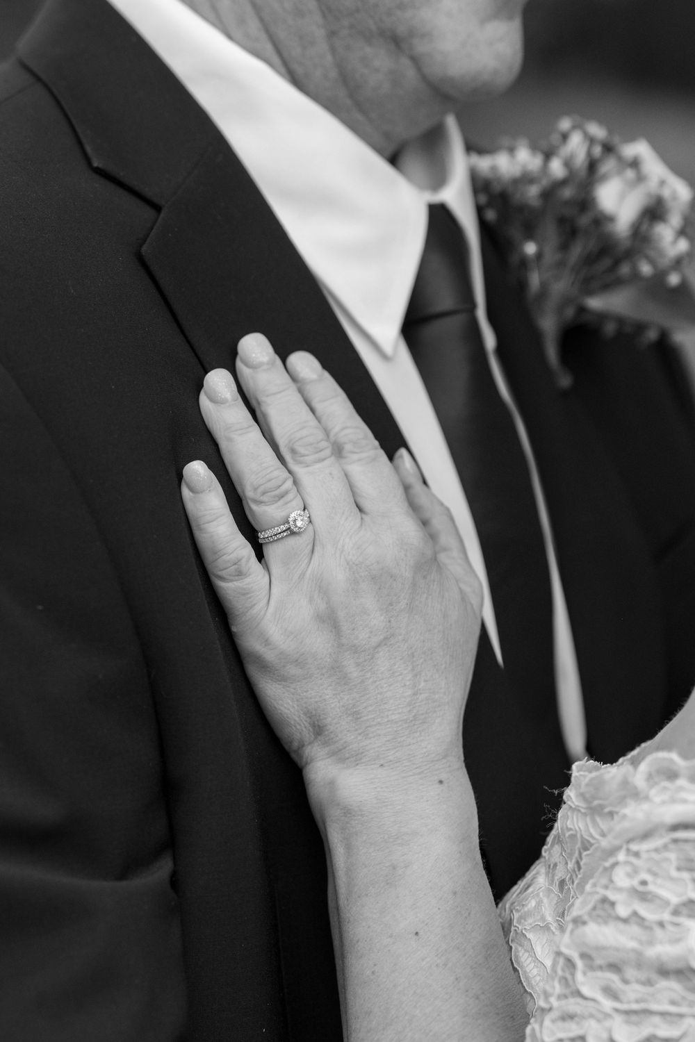 Black and white close-up of bride's hand with engagement ring on groom's suit.