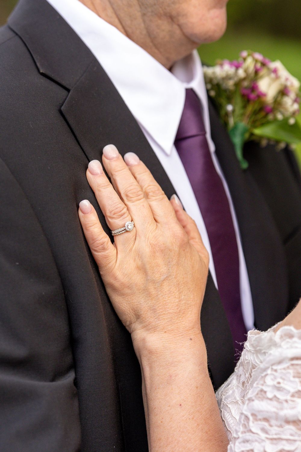 Bride's hand with diamond engagement ring resting on groom's black suit jacket at a wedding ceremony.