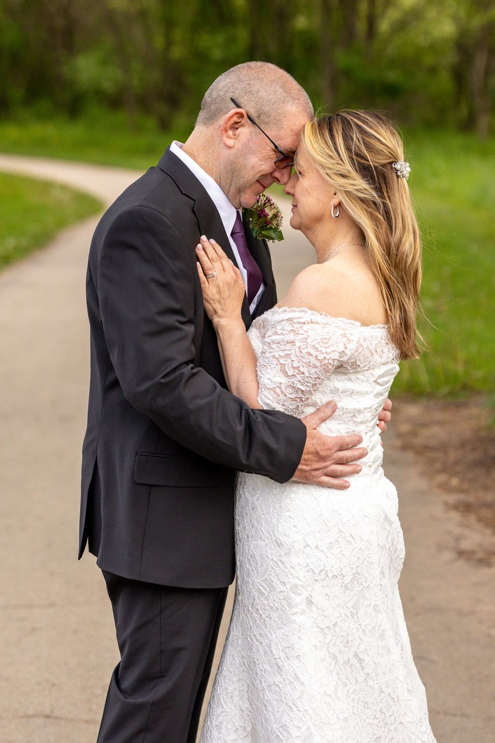 Bride and groom share an intimate forehead touch moment on a tree-lined path in their wedding attire.