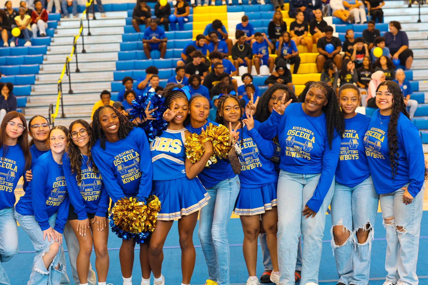 Cheerleaders and students in blue hoodies pose together in stadium bleachers at a school pep rally.