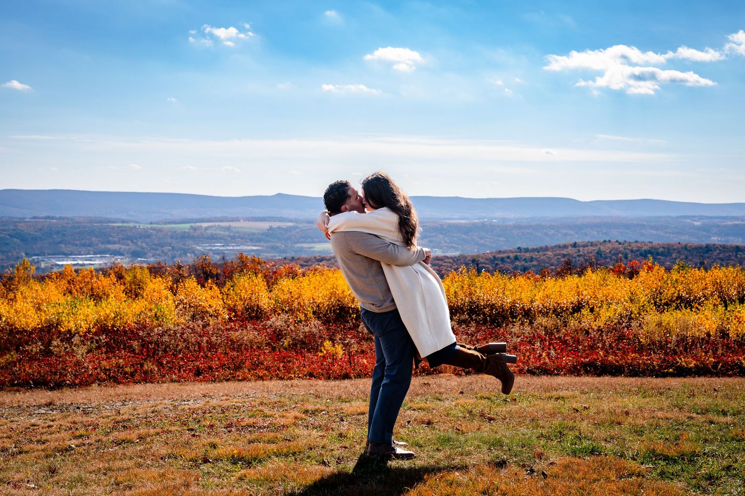 Couple embracing in autumn vineyard landscape with colorful fall foliage and rolling hills under blue sky.