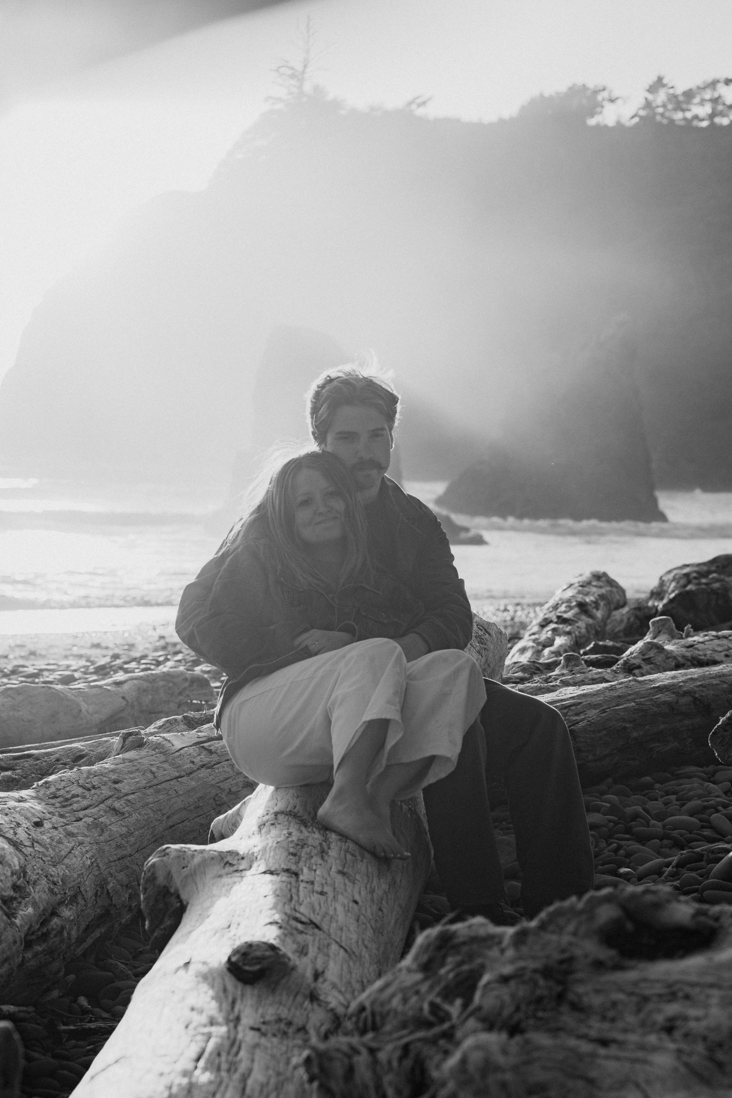 Couple embracing on driftwood log at misty beach, black and white romantic coastal photography.
