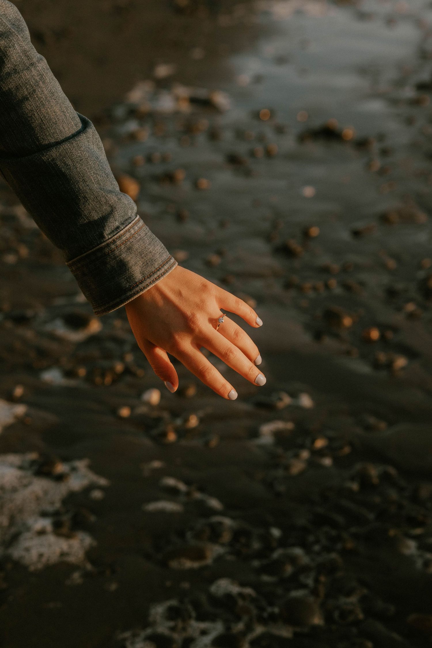 Two hands gently touching fingertips at golden sunset, one wearing a ring, set against a blurred coastal landscape.