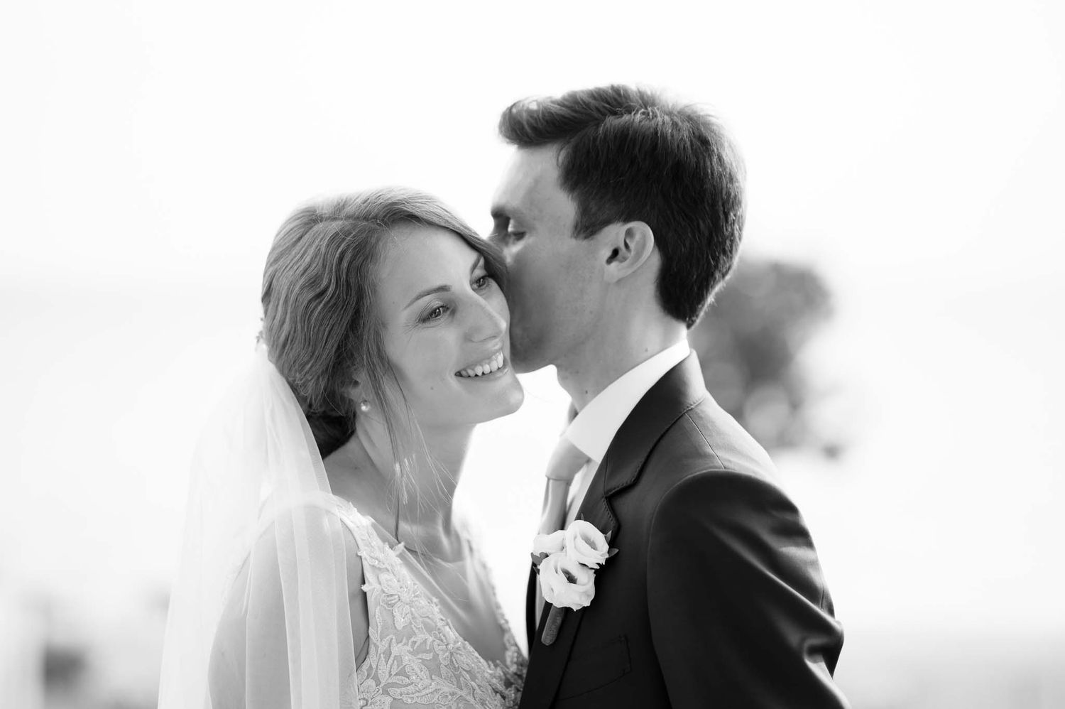 Black and white wedding photo of smiling bride in veil and groom kissing her forehead outdoors.