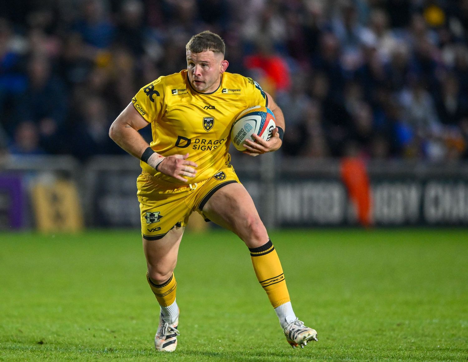 Rugby player in yellow kit runs with ball during evening match.