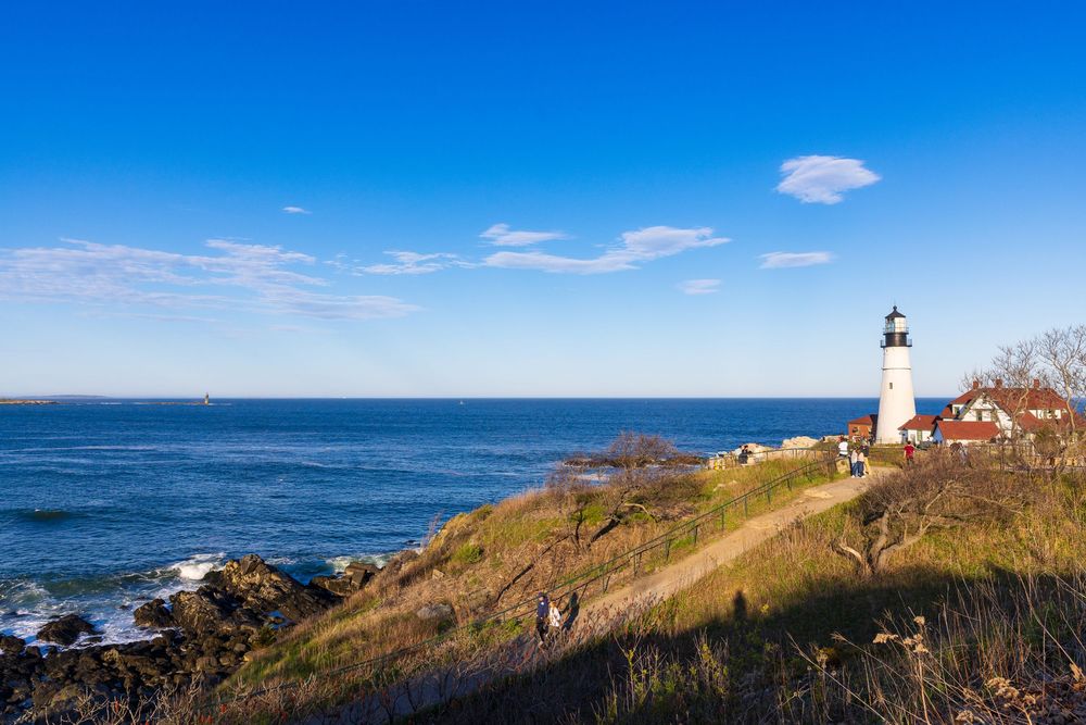 A white lighthouse stands on a rocky coastal cliff overlooking the blue ocean under a clear sky.