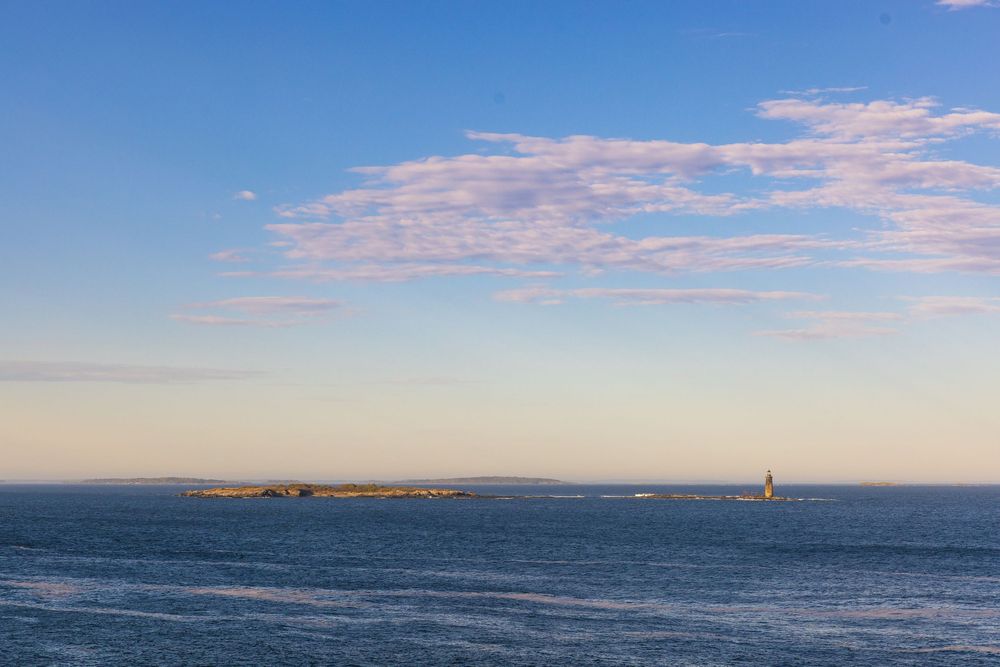 Panoramic ocean view with a small island and lighthouse under a blue sky with wispy clouds at golden hour.
