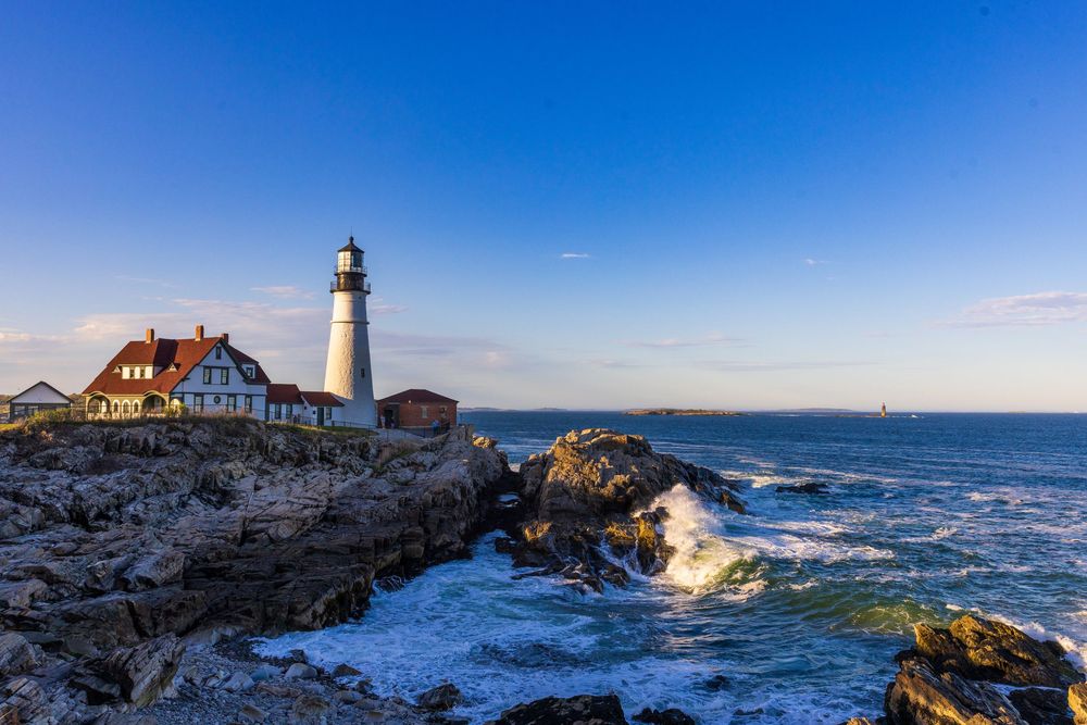 White lighthouse on rocky Maine coastline with crashing waves under a clear blue sky at golden hour.
