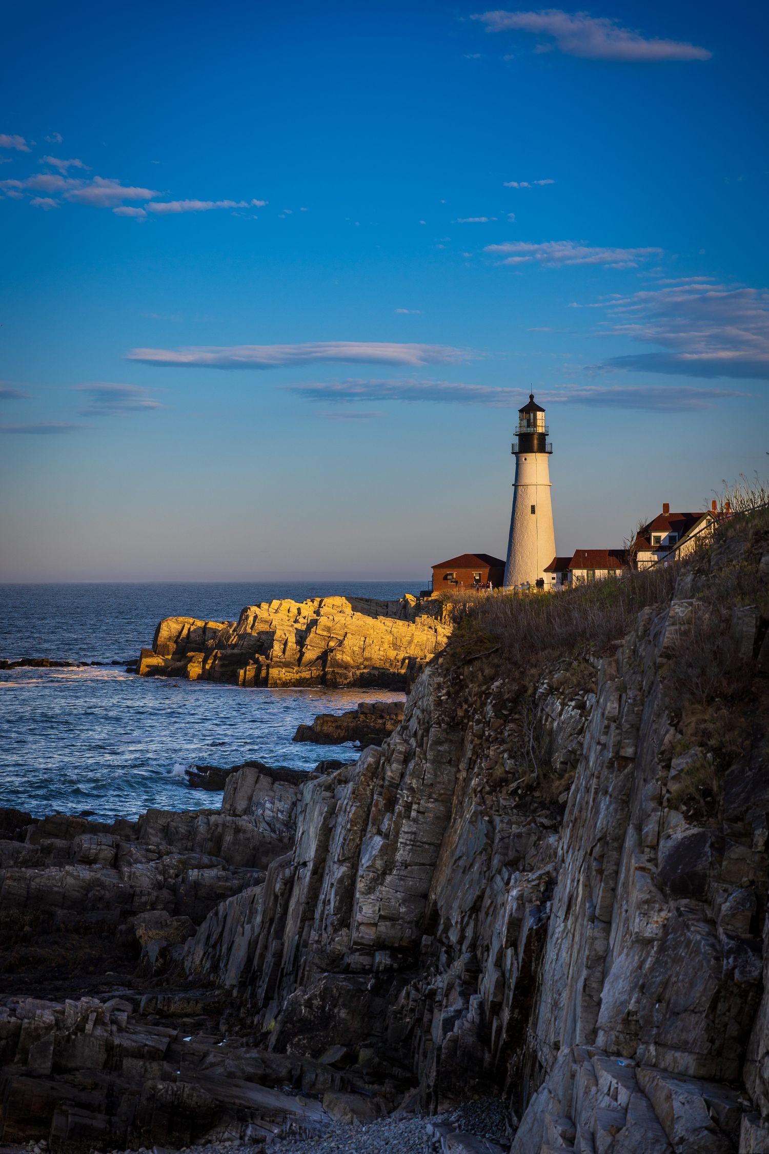 White lighthouse on rocky Maine coastline at golden hour with blue sky and ocean waves below.