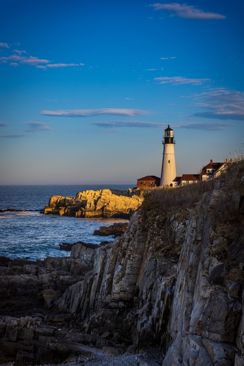 White lighthouse on rocky Maine coastline at golden hour with blue sky and ocean waves below.
