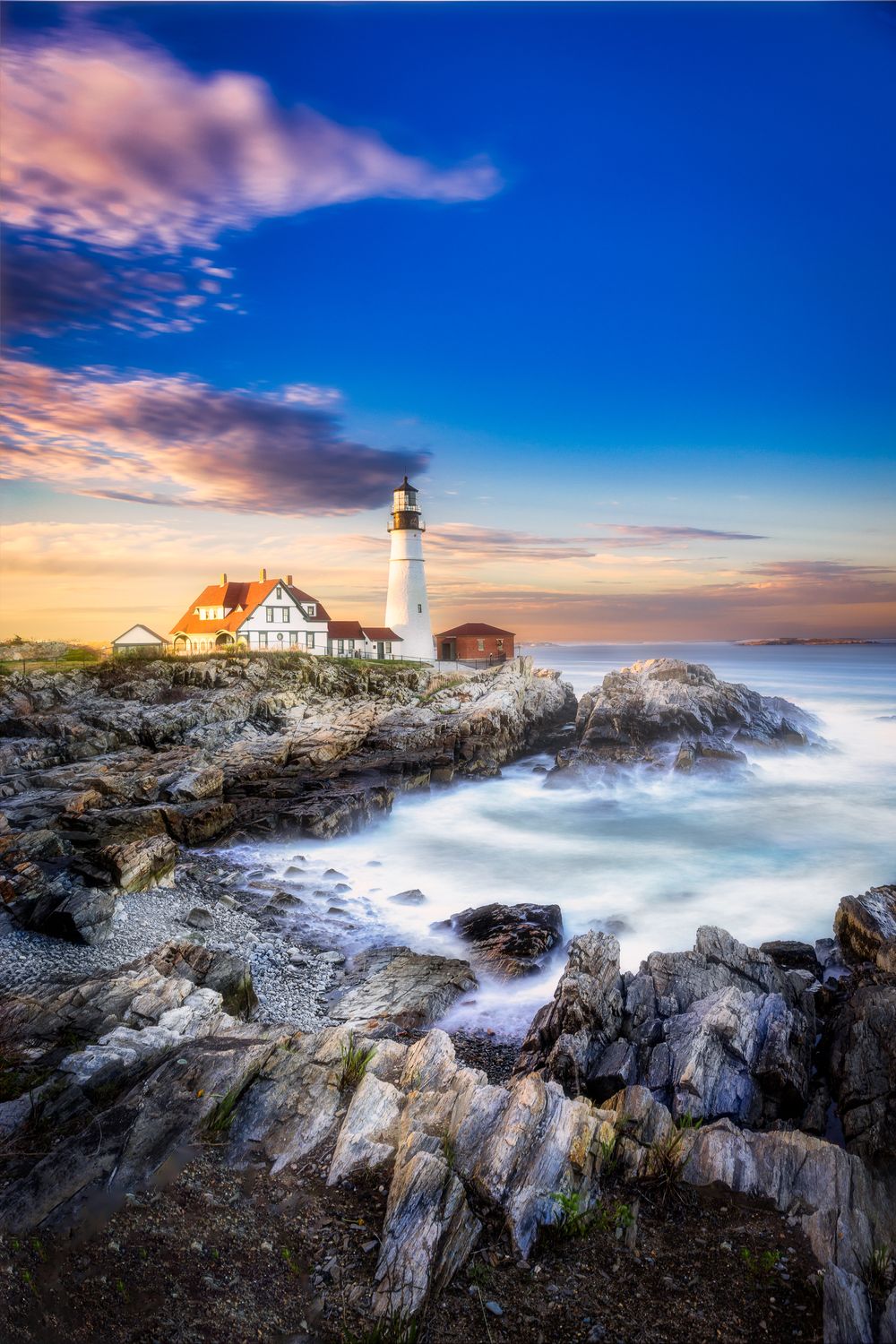 Portland Head Lighthouse on rocky Maine coast at sunset with silky waves and dramatic blue sky.