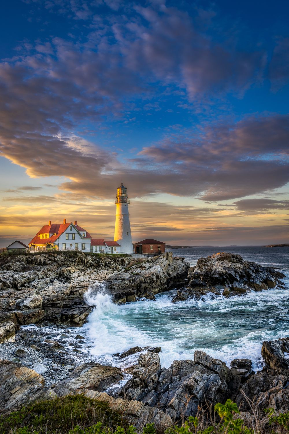 Portland Head Lighthouse at sunset with crashing waves on rocky Maine coastline under dramatic blue sky.